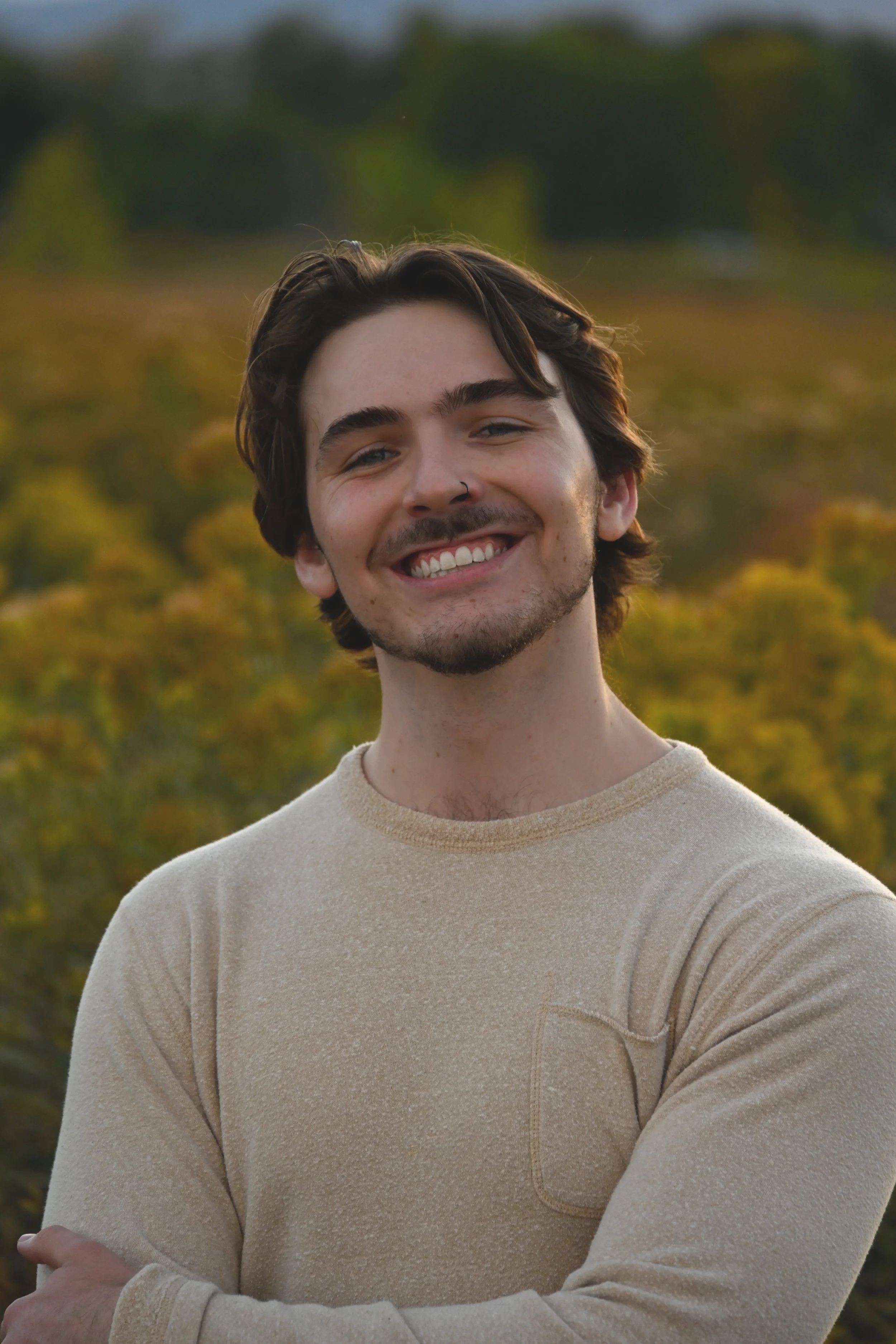 A young man with dark hair, a nose ring, and a beard smiling outdoors with a blurred green and yellow landscape in the background.