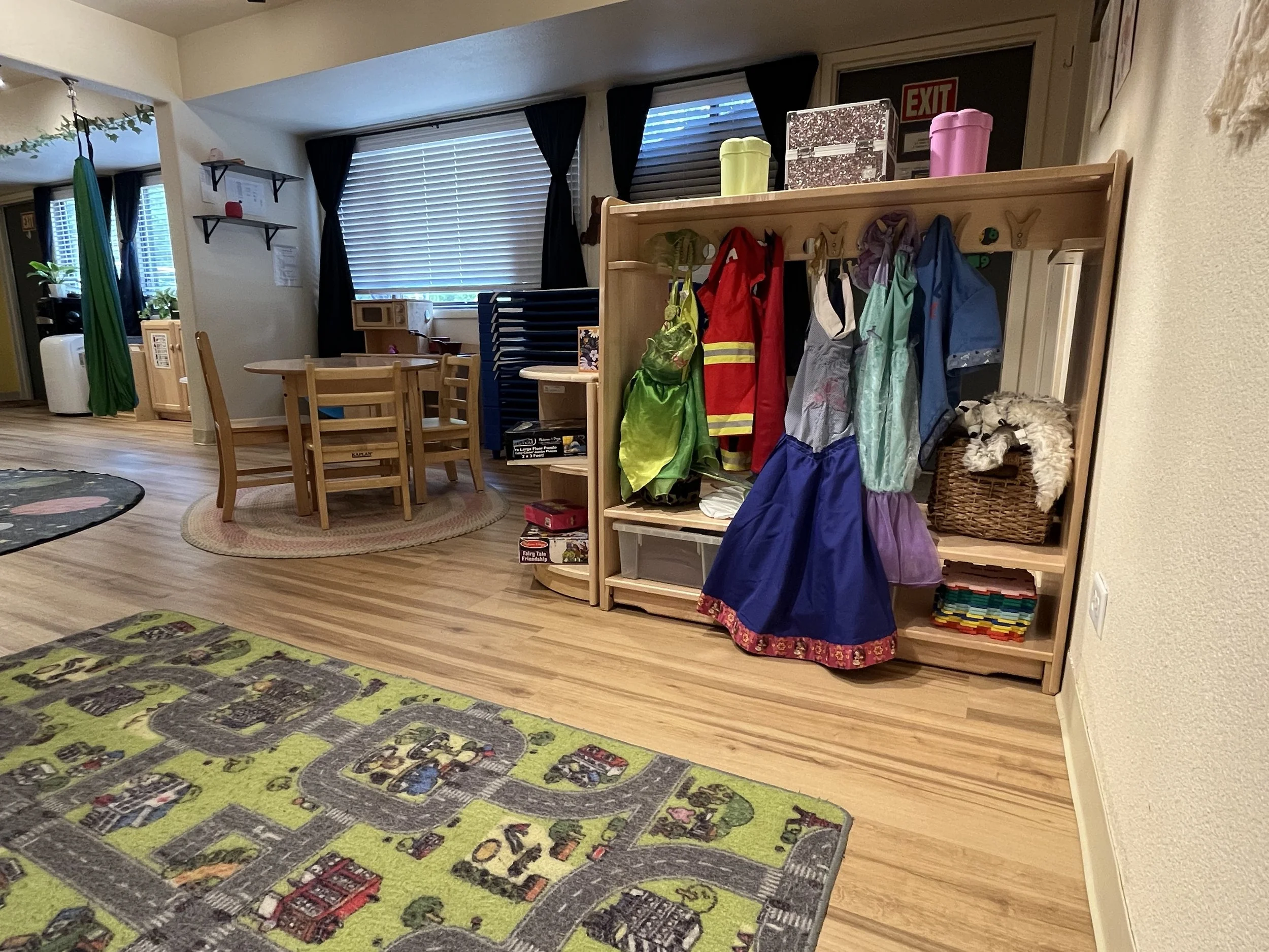 Children's play area with a colorful wooden coat rack holding costumes and accessories, a basket with stuffed animals, a wooden table with chairs, a play rug with roads and buildings, and a corner with shelves and toys, in a cozy room with wood flooring and window with blinds.
