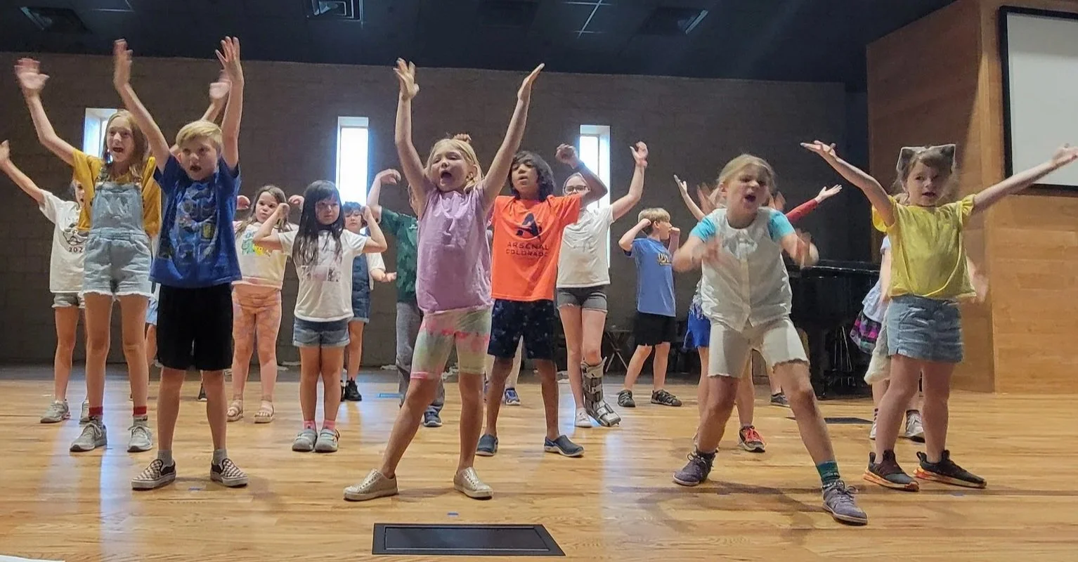Children in animal and farm-themed costumes perform on stage during a school play. Kids are dressed as cows, pigs, cows, and other animals and are dancing or striking poses.