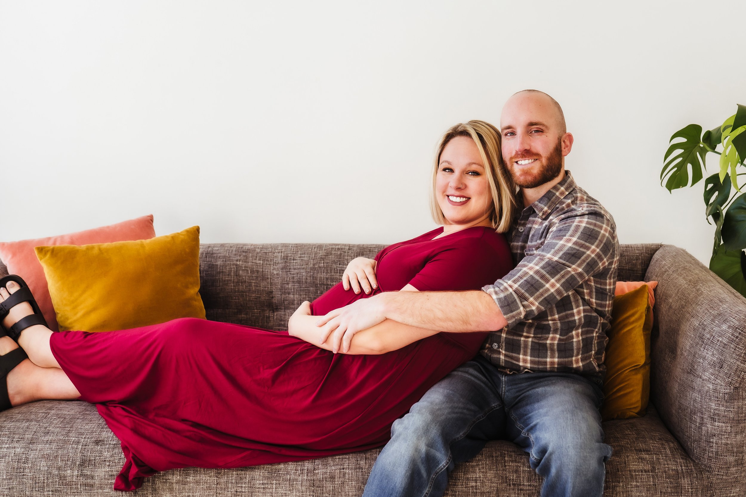 Pregnant couple cuddle on the couch smiling at photographer