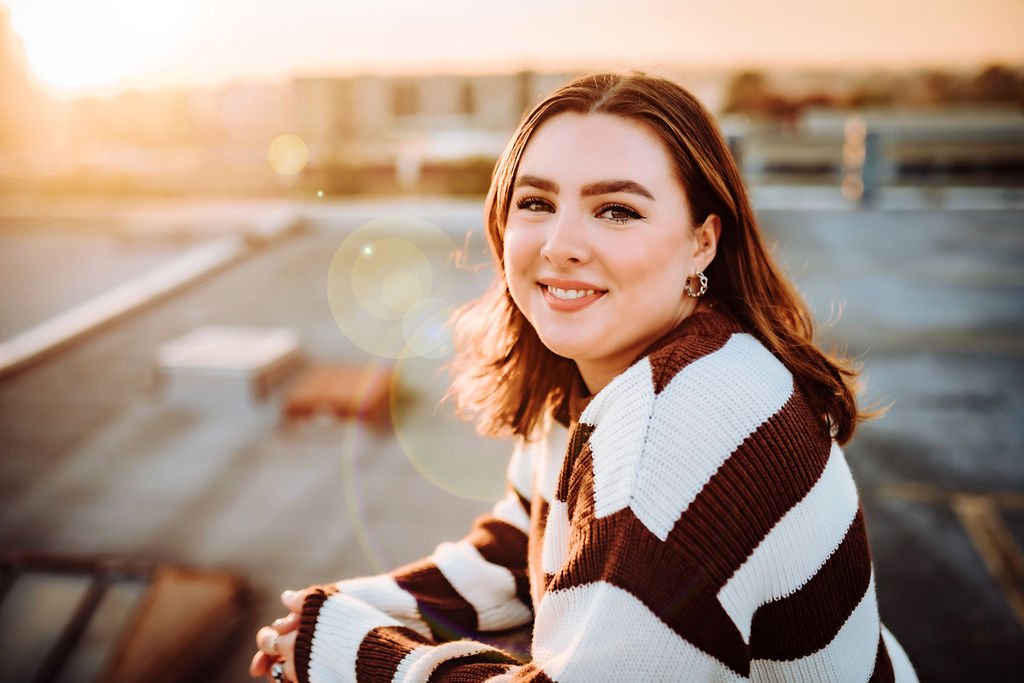 At sunset senior girl leaning over wall with sun flare looking at camera