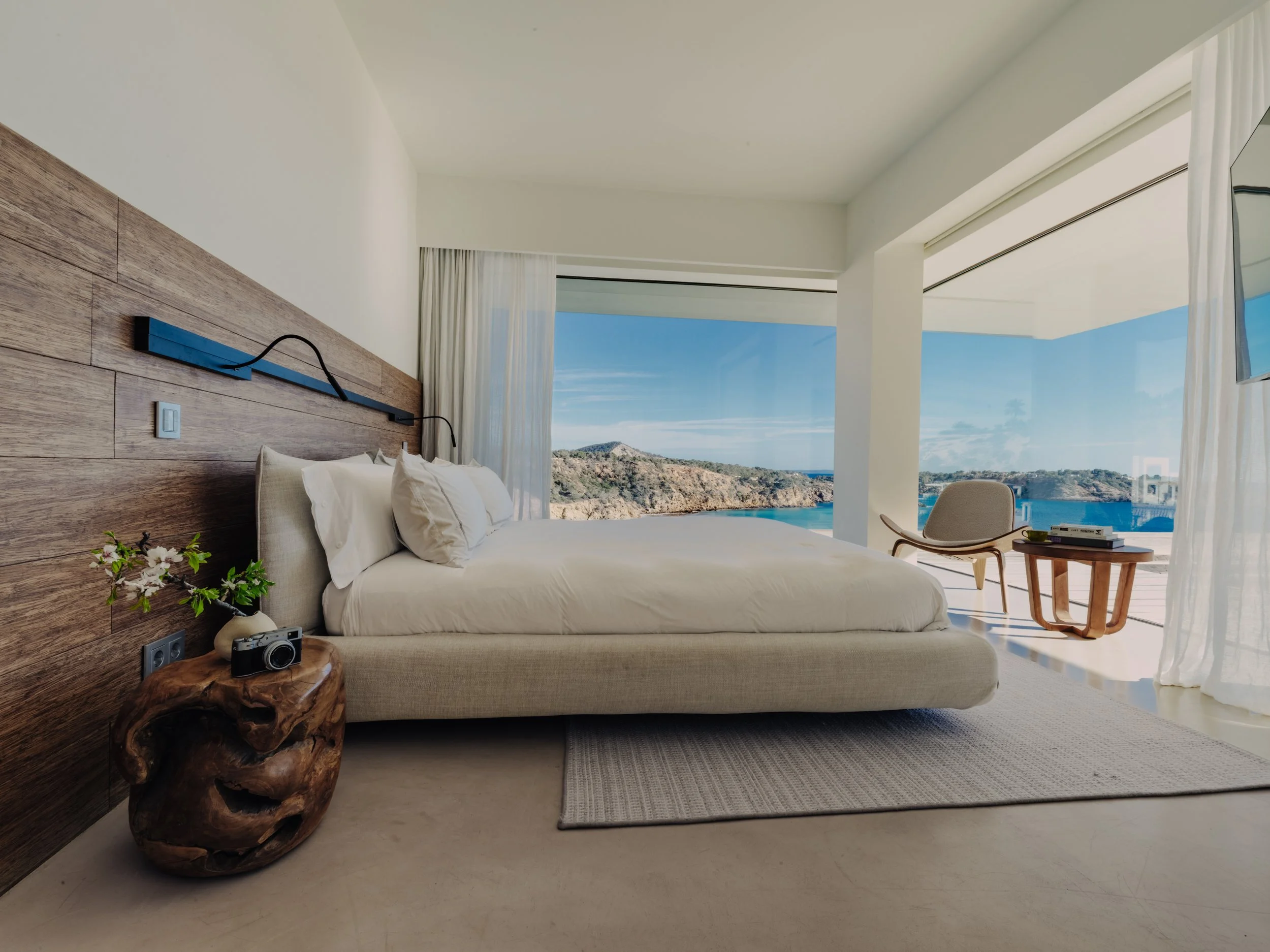 Modern bedroom with a large bed, a wooden nightstand with a camera and plant, white curtains, and a view of the ocean and mountains through glass doors.