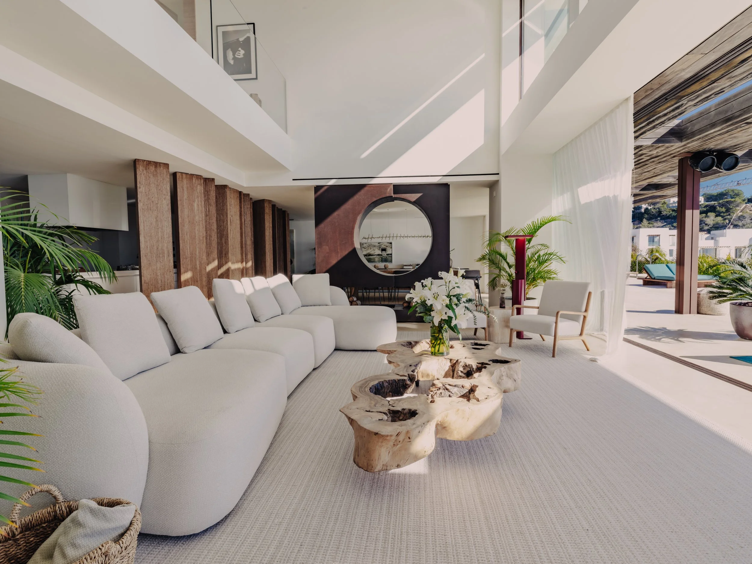 Modern living room with a large white sectional sofa, wooden coffee table, potted plants, and a round mirror on the wall, with sunlight streaming through large windows.