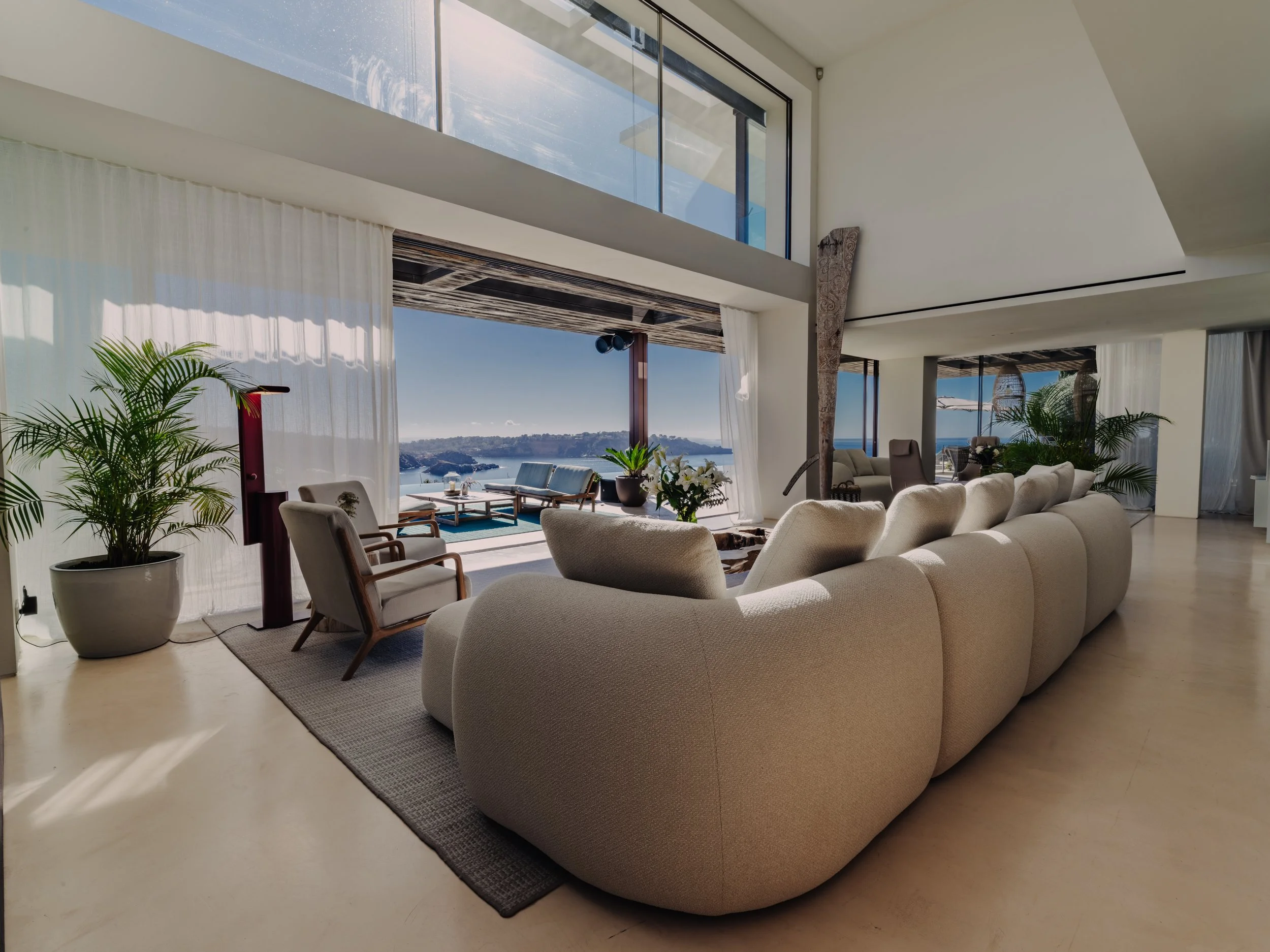 Modern living room with large beige sectional sofa, green plants, a rug, and sliding glass doors leading to an outdoor patio with ocean views.