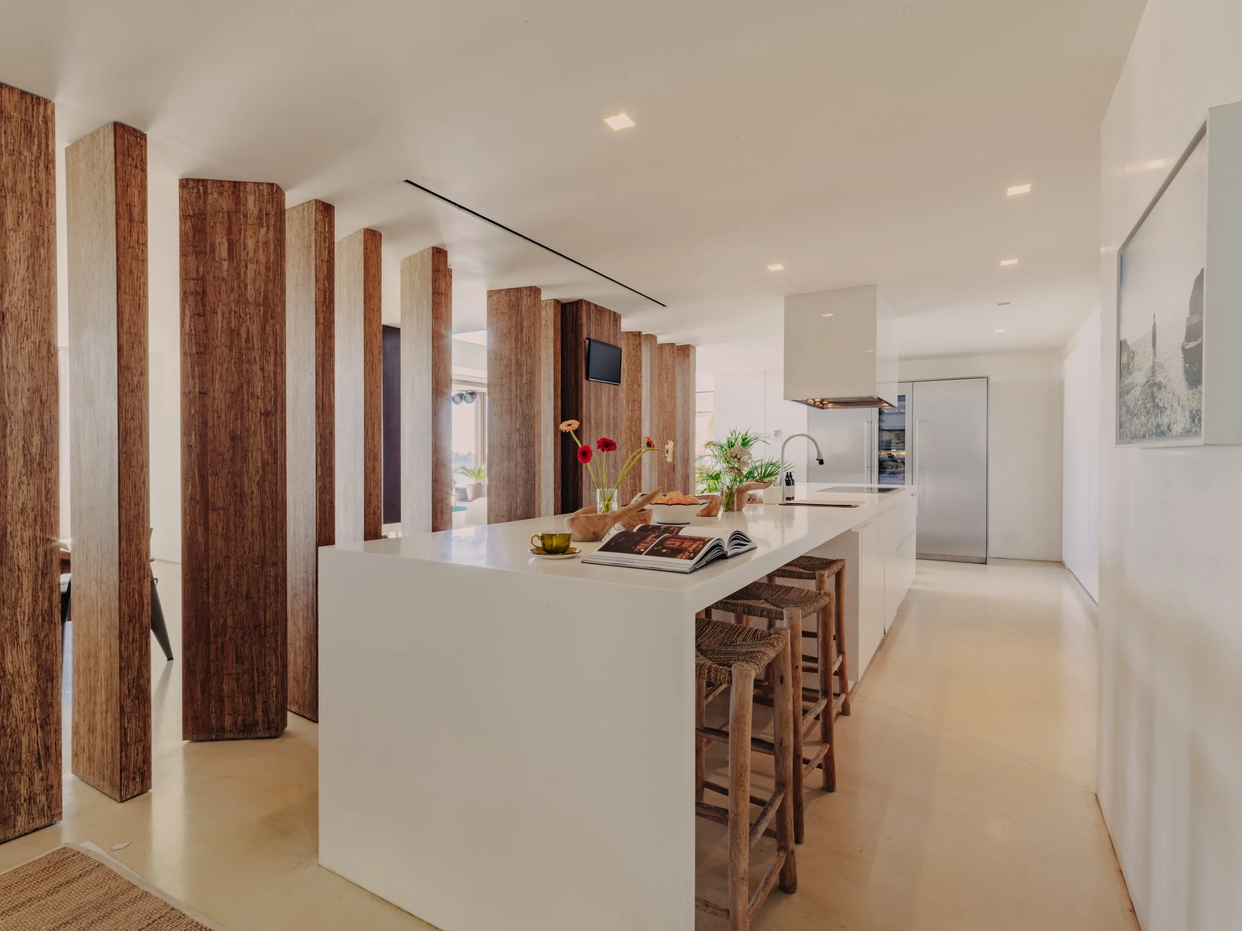 Modern kitchen with white island counter, wooden stools, and wooden partition walls, with flowers and books on the counter and a stainless steel refrigerator in the background.