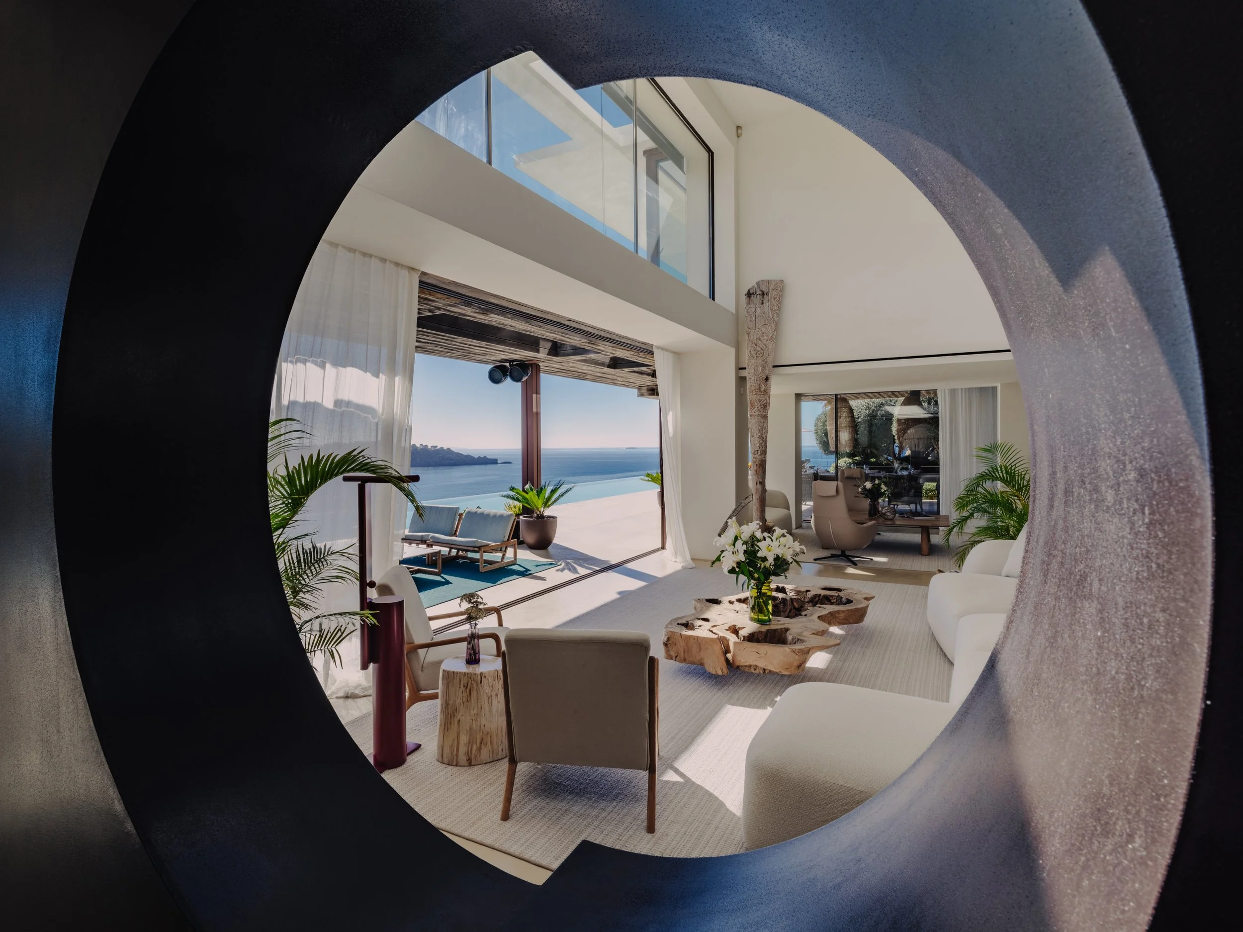 Modern living room with sitting area and ocean view, seen through a circular opening in a wall.
