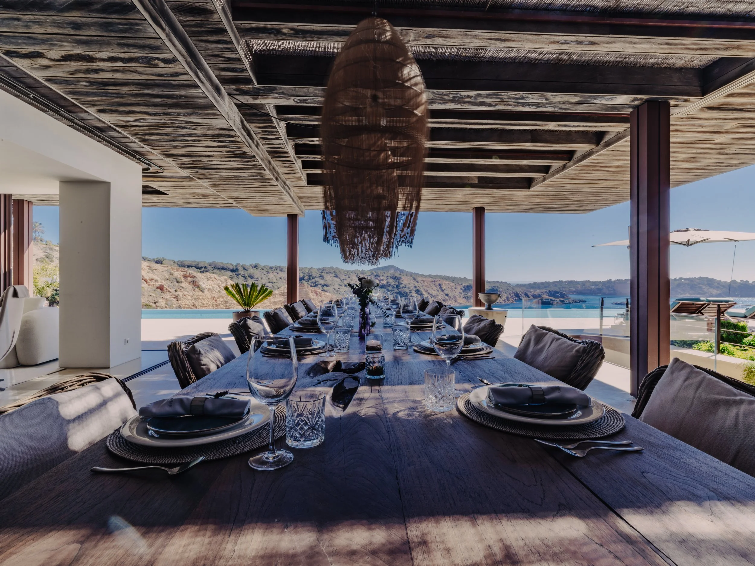 A spacious outdoor dining area with a long wooden table set with plates, glasses, and napkins. The area overlooks a scenic body of water and hills under a clear blue sky. There are cushioned chairs, a large tropical plant, and a unique hanging lamp w