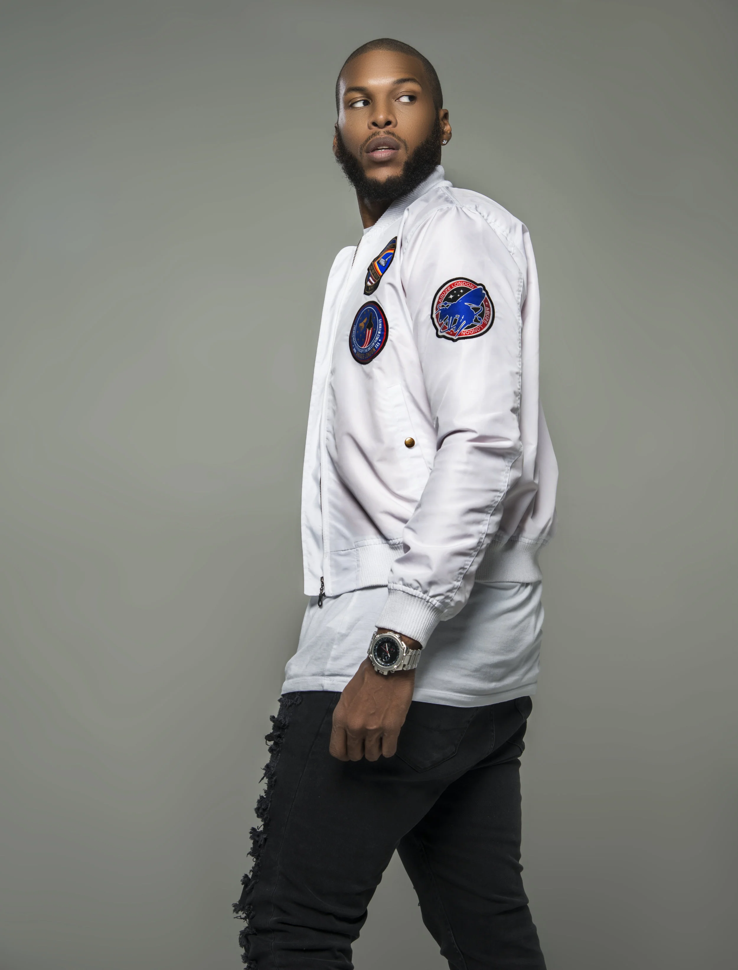 Young man with a beard and earrings wearing a white NASA jacket and black ripped jeans, standing against a plain gray background.