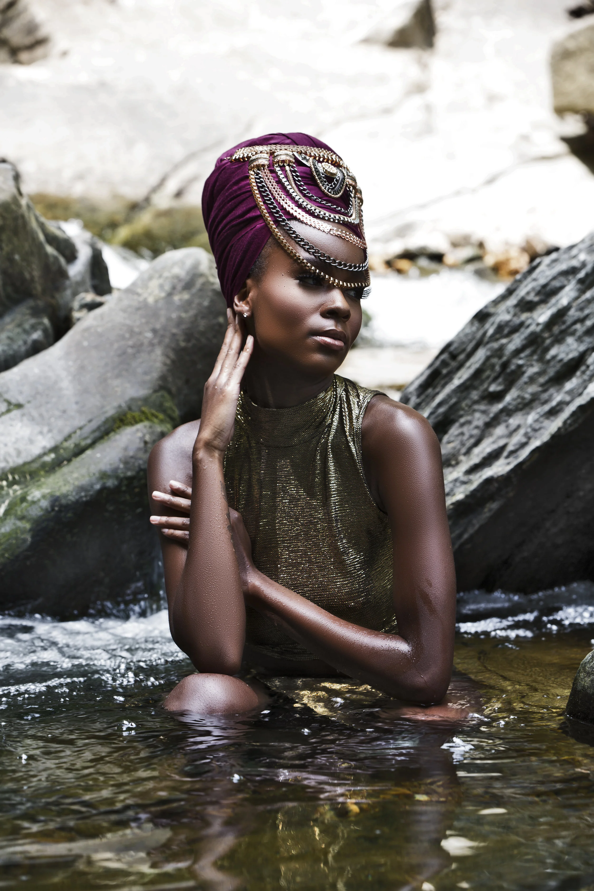 A woman with dark skin, gold metallic dress, and an elaborate purple headwrap adorned with silver chains and gemstones, sitting in a shallow stream surrounded by rocks and natural scenery.