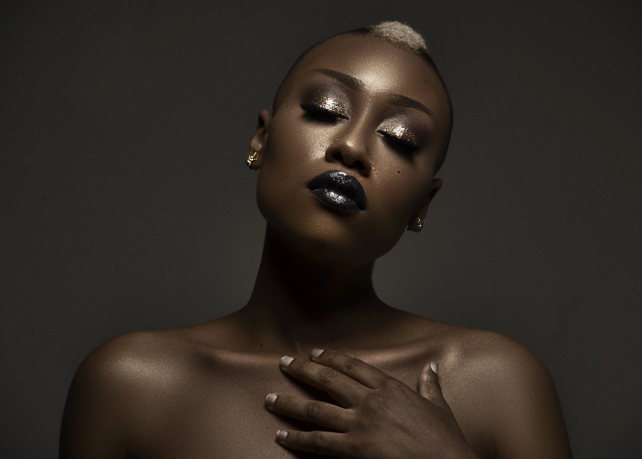 Close-up of a woman with dark skin and short blonde hair, makeup with shimmering eyeshadow and black lipstick, displaying her bare shoulders and a hand resting on her chest against a dark background.
