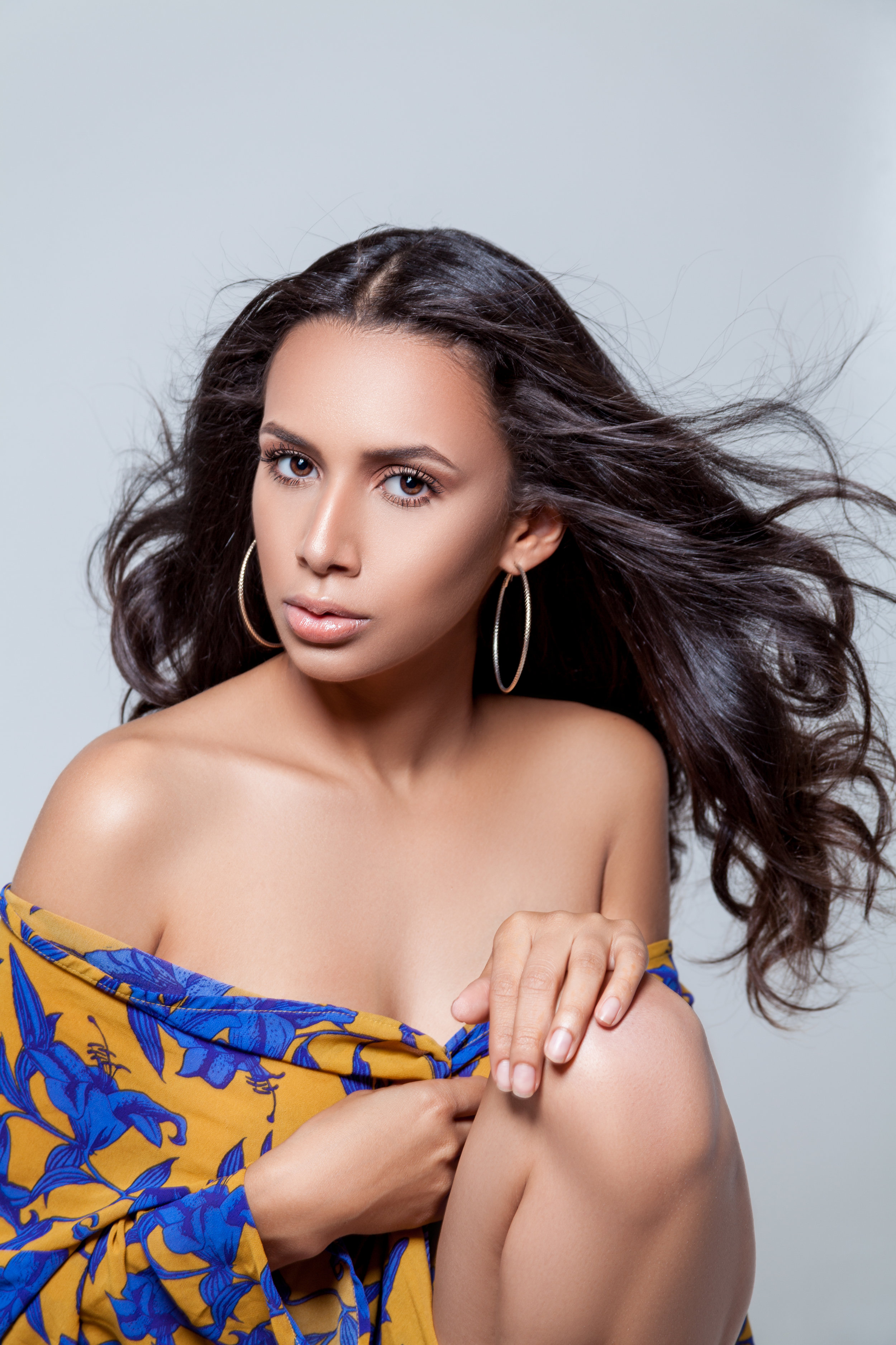A woman with long dark hair, wearing hoop earrings, a yellow and blue patterned off-the-shoulder top, and minimal makeup, posing against a plain light gray background.