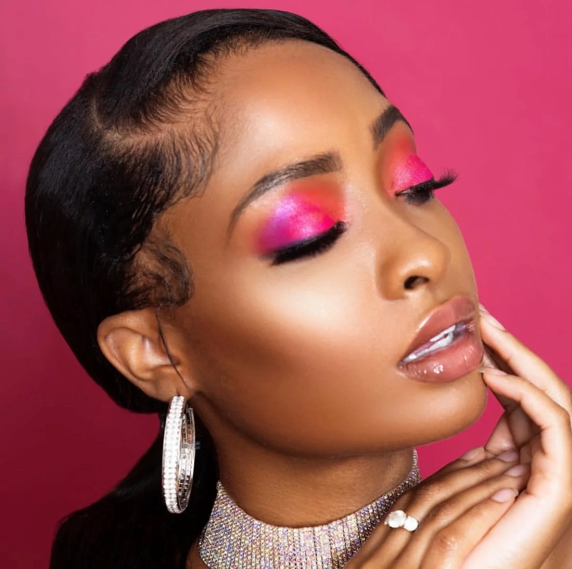 Close-up of a woman with dark hair styled close to her scalp, wearing pink and purple eyeshadow, long eyelashes, glossy lips, and jewelry including large hoop earrings, a diamond necklace, and a ring, against a pink background.