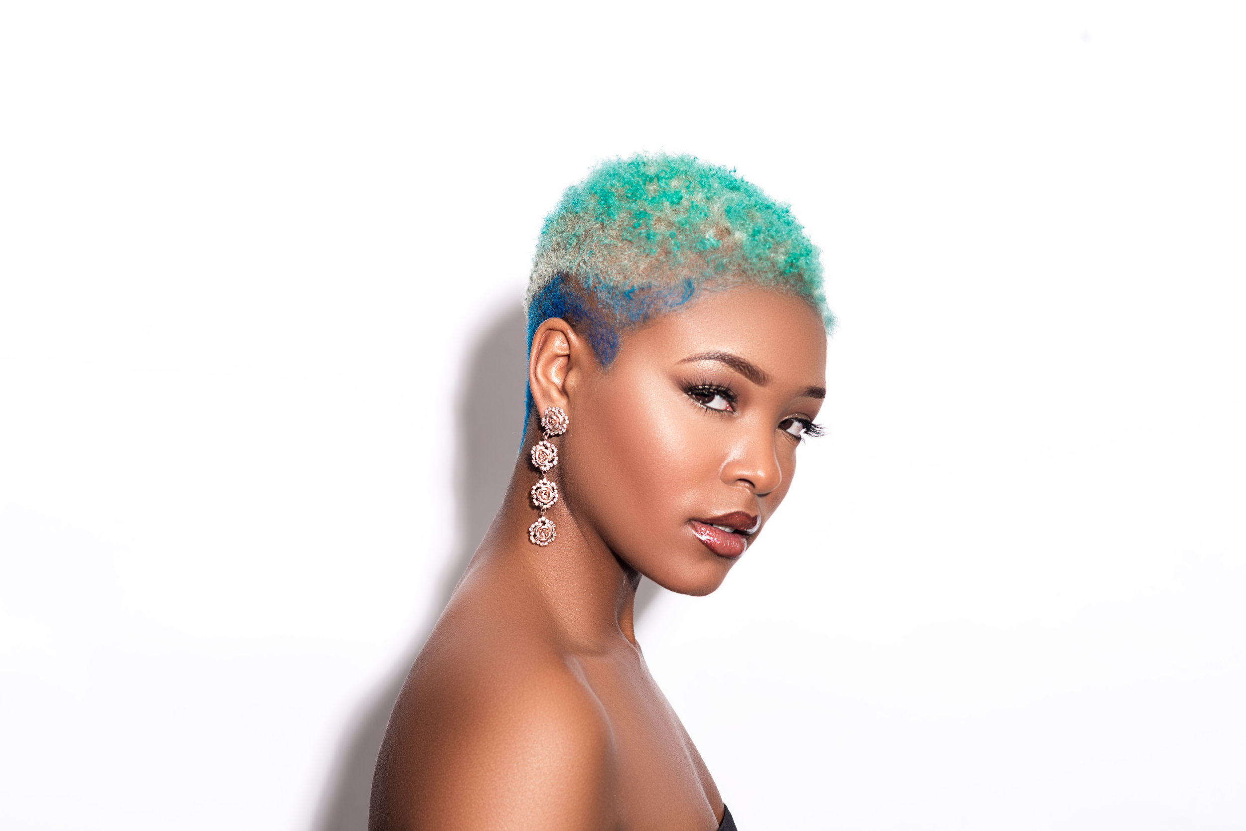 A woman with short, curly hair dyed in pastel shades of green, blue, and blonde, wearing large, ornate earrings, and looking confidently at the camera against a plain gray background.