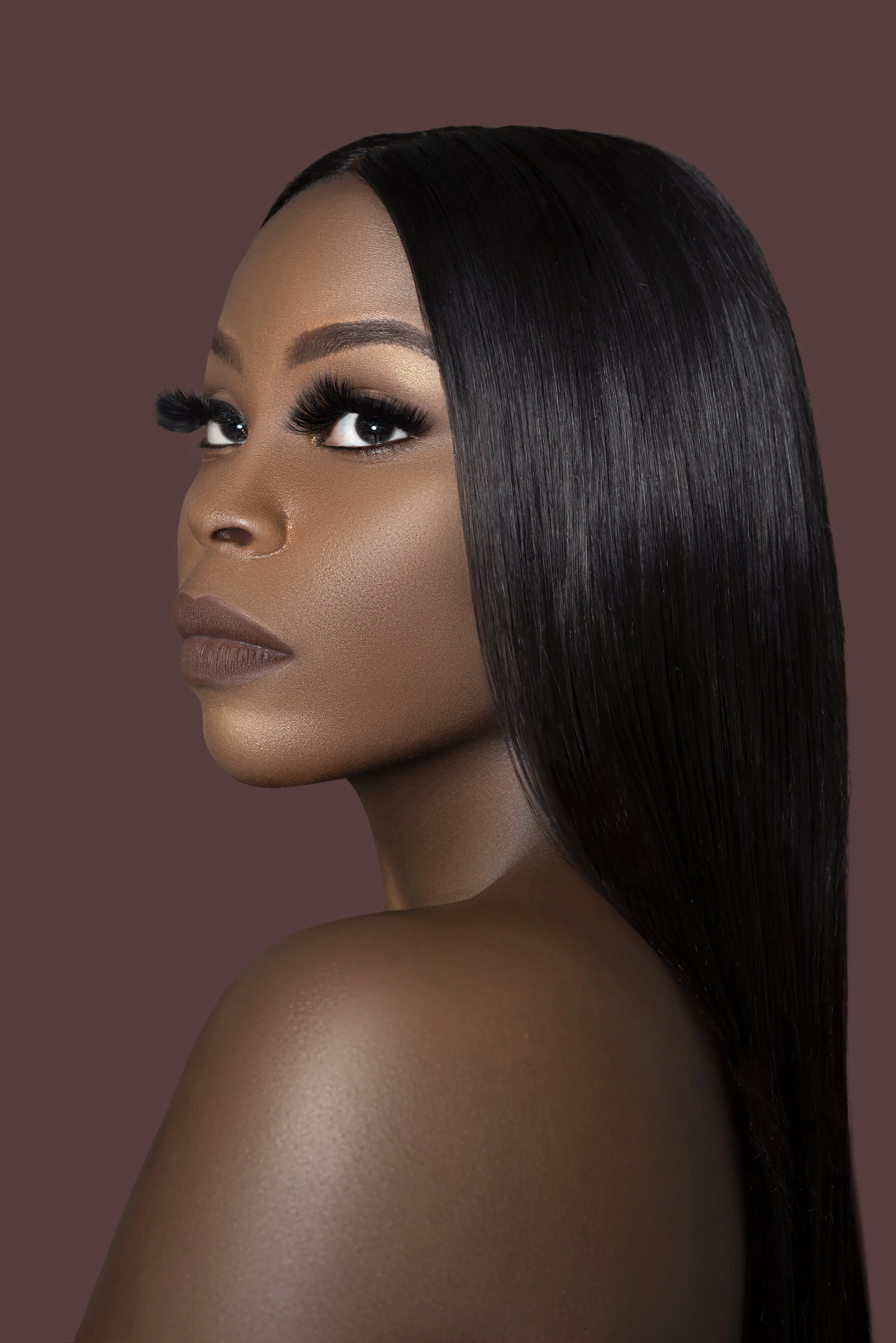 Close-up of a woman with smooth, straight black hair, light brown makeup, full lips, and long eyelashes, posing against a muted brown background.