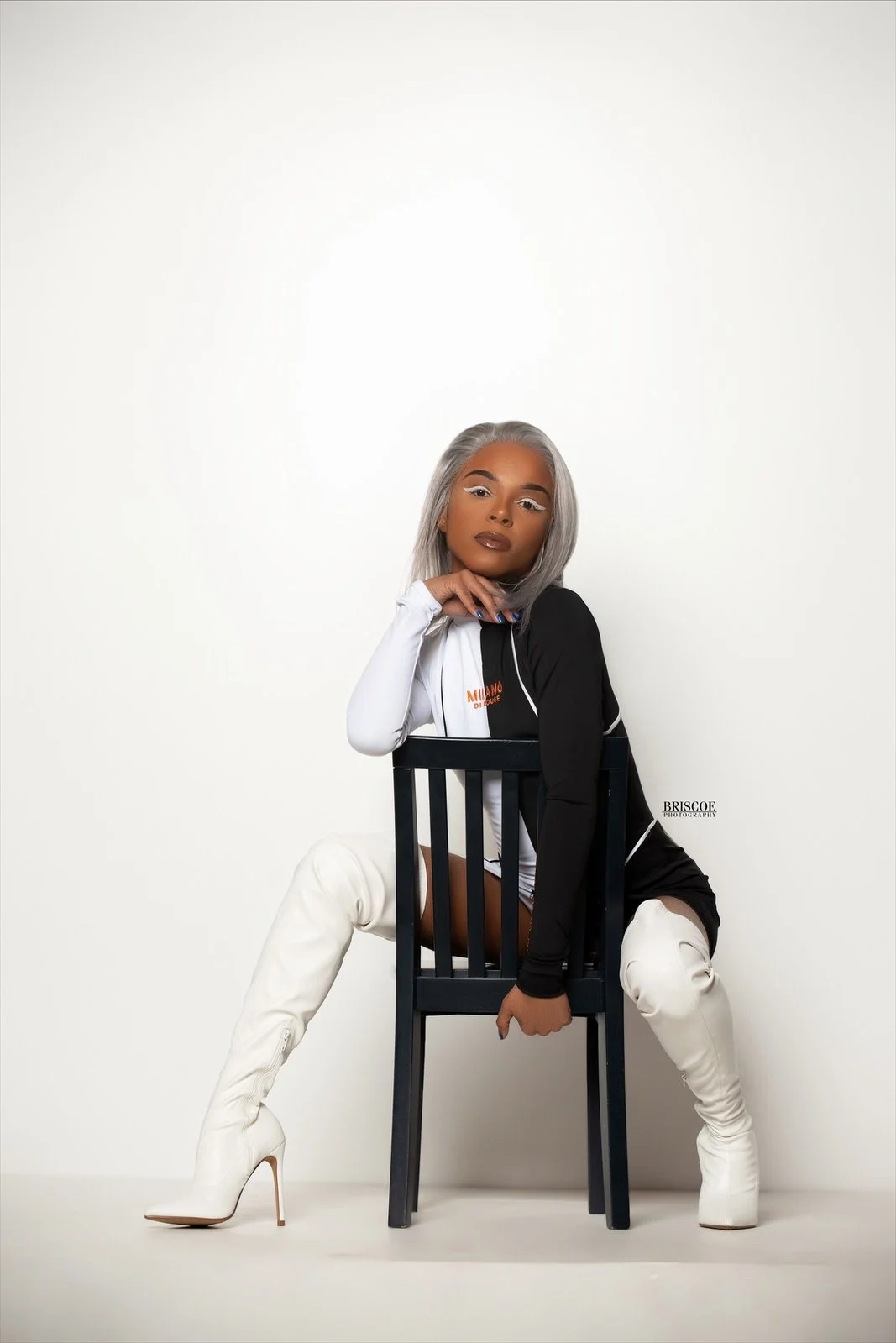 A woman with silver hair, wearing a black and white outfit and white thigh-high boots, poses sitting on a black chair against a plain white background.
