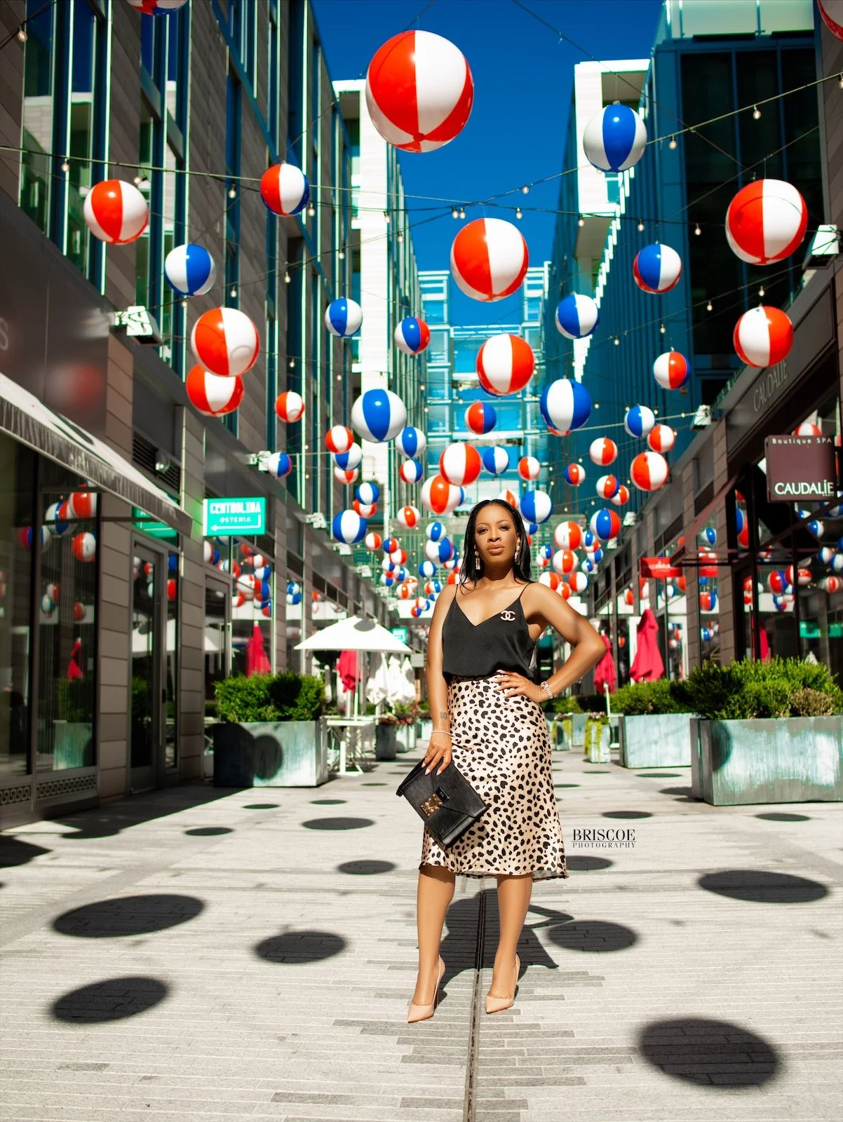 A woman posing in an outdoor shopping district decorated with red, white, and blue striped lanterns hanging overhead on a sunny day.
