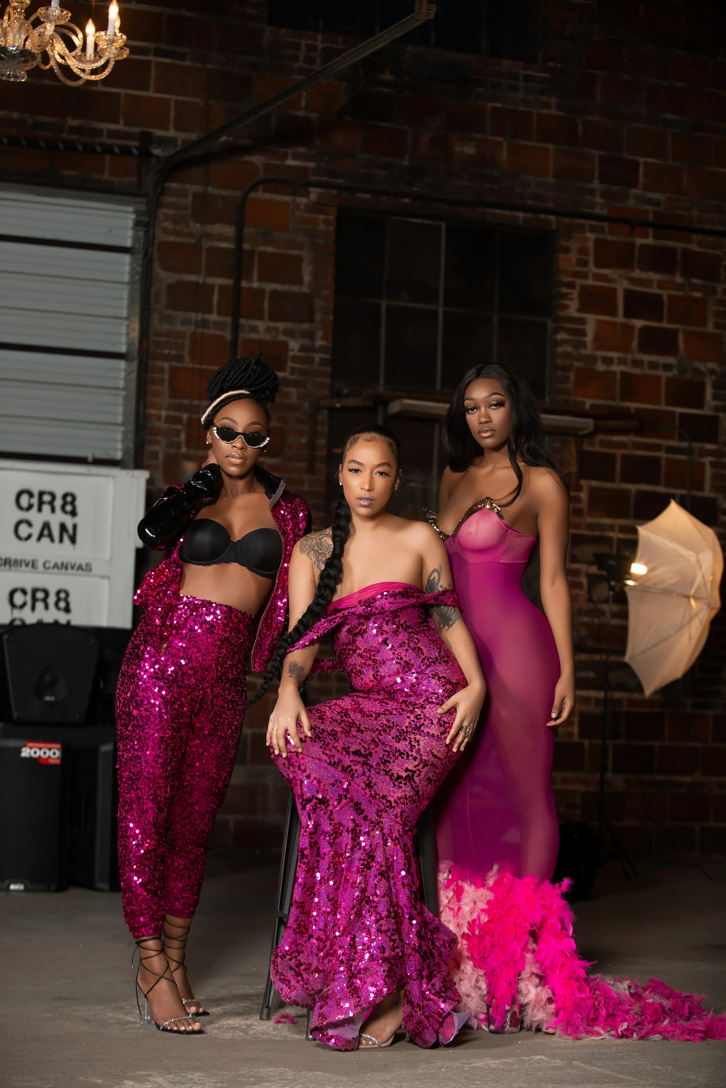 Three women are dressed in glamorous pink and purple outfits, posing indoors against a brick wall. There is a chandelier hanging above them and a vintage photography umbrella to the right. The woman on the left is wearing a sequined pink jacket and m