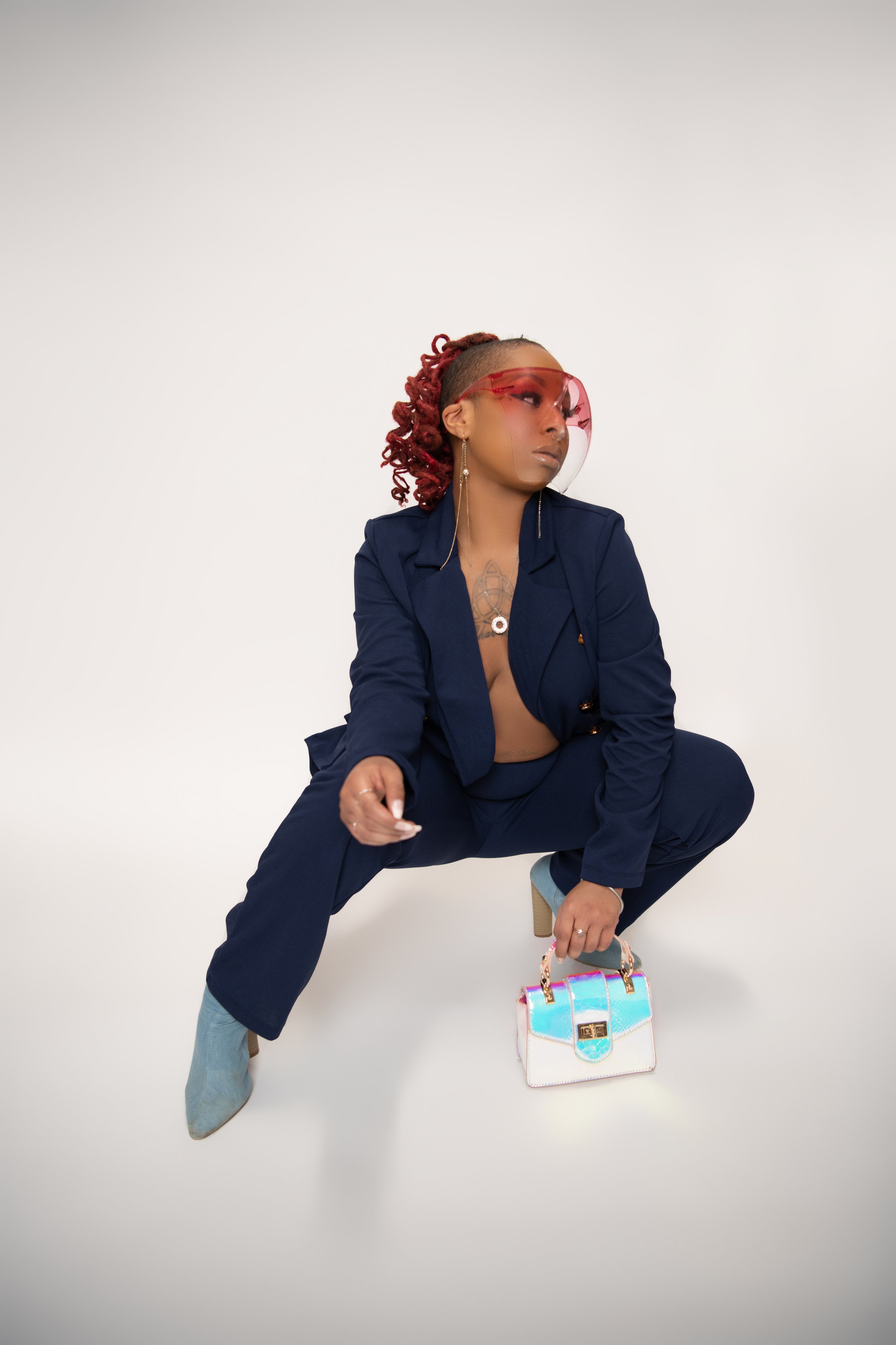 Fashion-forward woman with red dreadlocks, oversized red visor sunglasses, navy blazer and pants, holding a colorful holographic handbag, posing against a plain white backdrop.