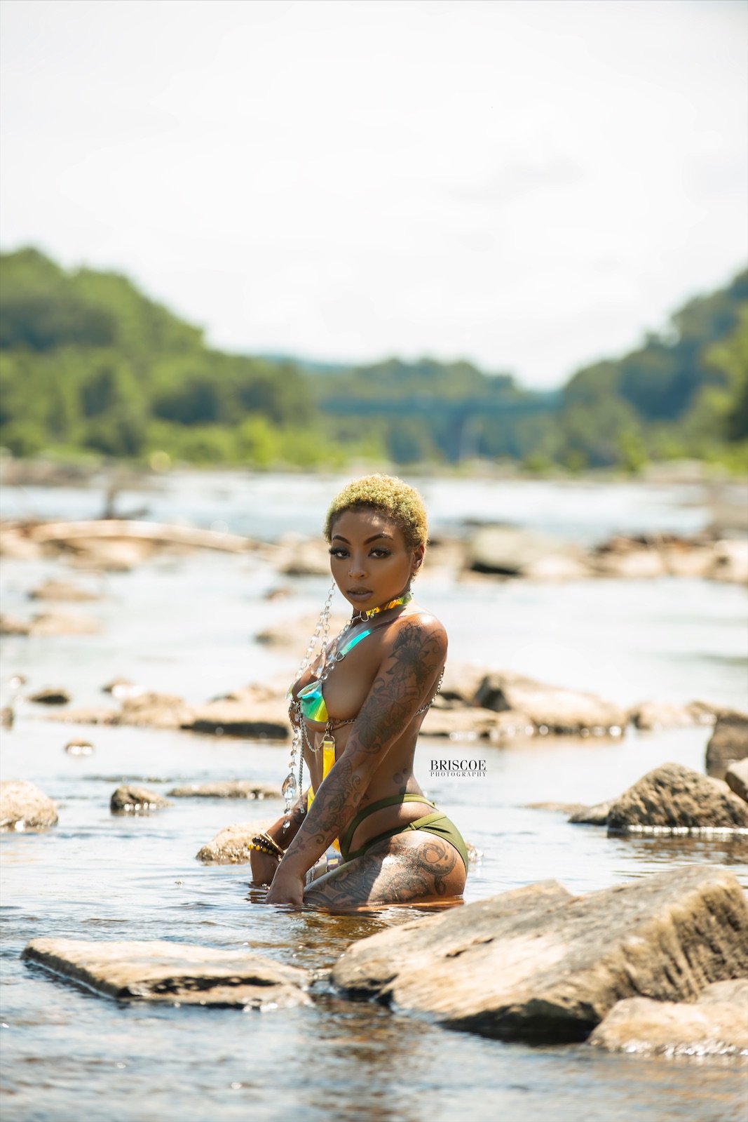 A woman with short blonde hair and tattoos is kneeling in a river, wearing swimwear and jewelry, with a natural landscape of trees and rocks in the background.