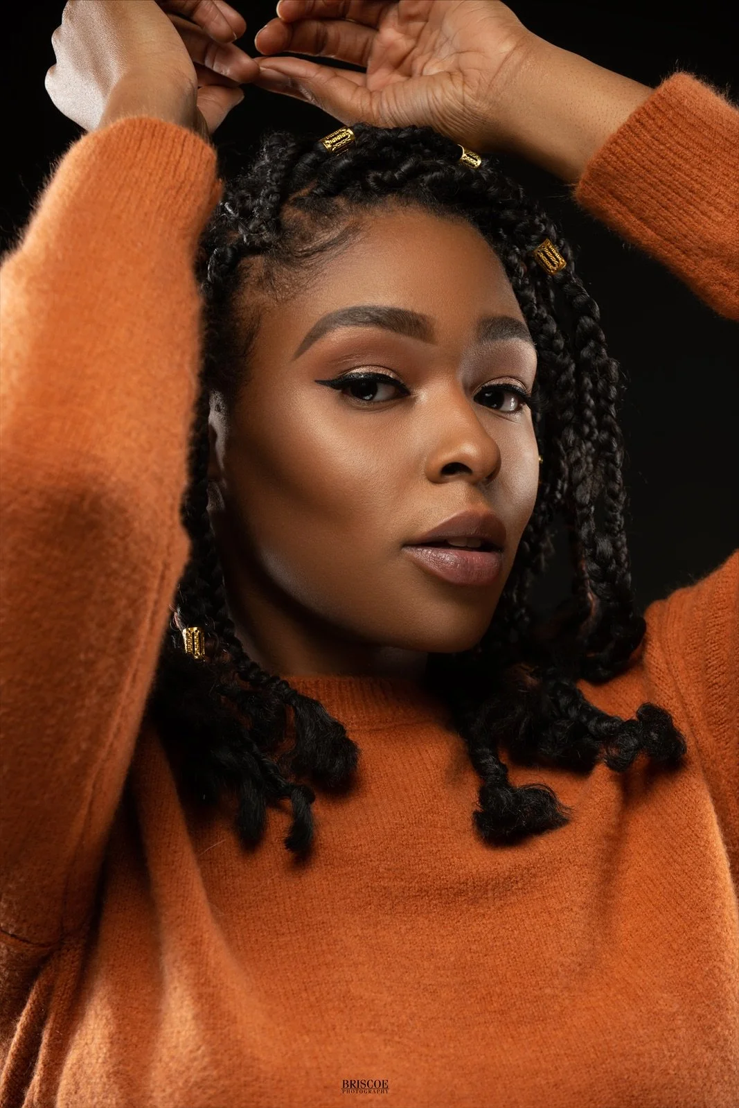 Close-up portrait of a woman with dark skin, braided hair with gold beads, bold eyeliner, wearing a rust-colored sweater, posing with hands above her head against a black background.