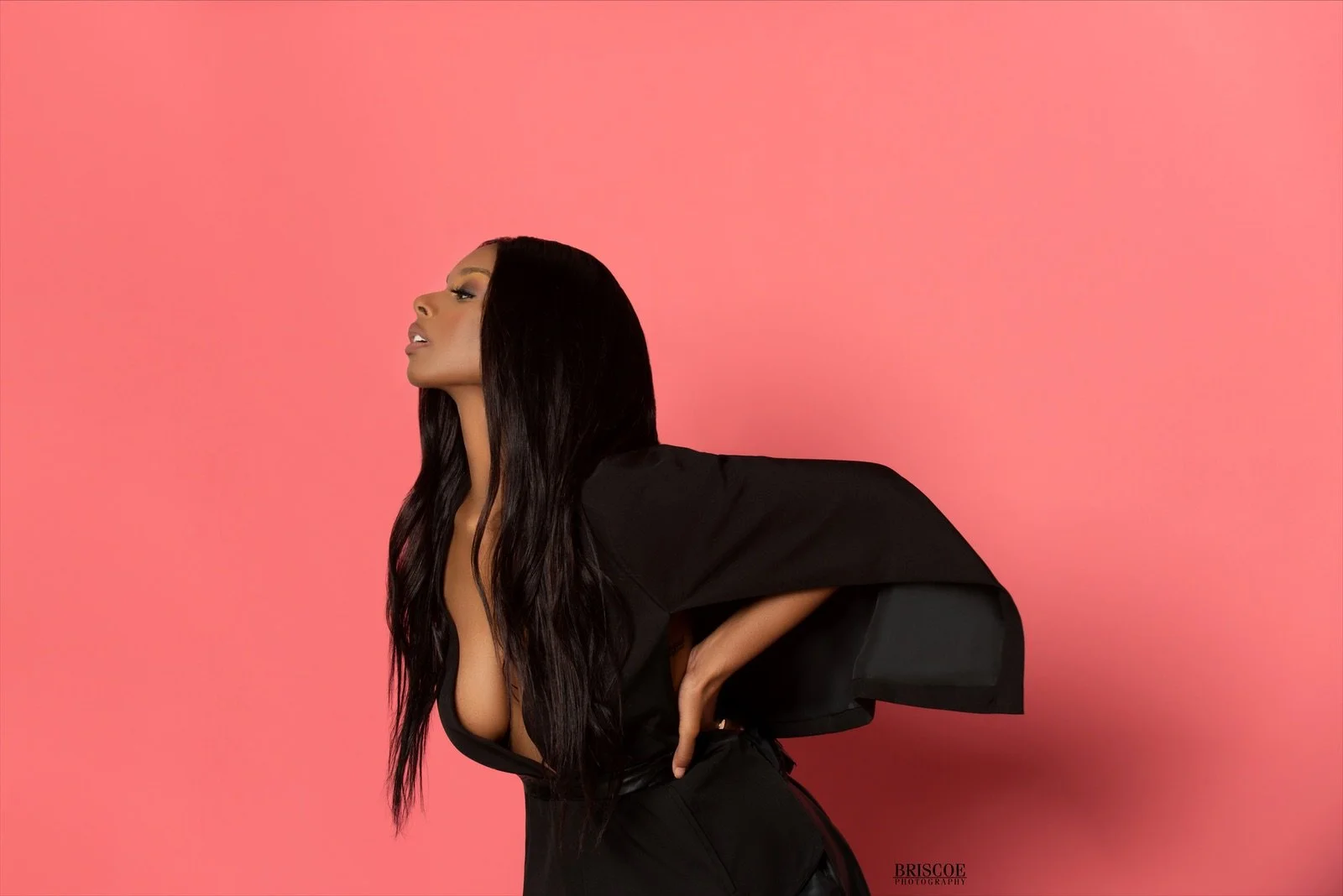 A woman with long black hair posing against a pink background. She is wearing a black top with a deep neckline and has her hand on her hip, with her head turned to the side.