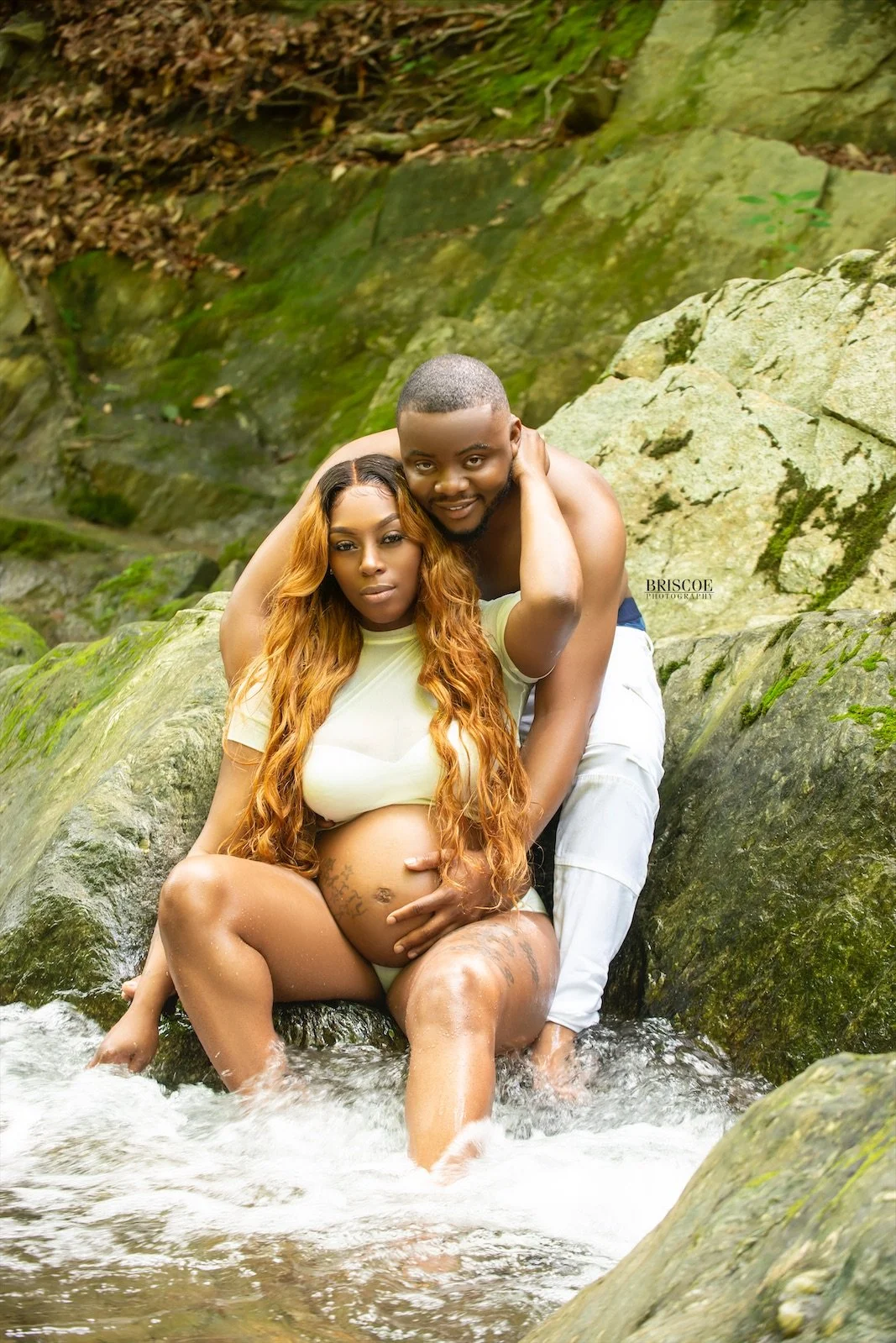 A pregnant woman with long, wavy hair and a man with short hair posing in a stream with rocks and moss in the background. The woman is sitting on a rock with her legs in the water, and the man is crouching behind her, both touching her belly, with the woman looking directly at the camera and the man smiling.