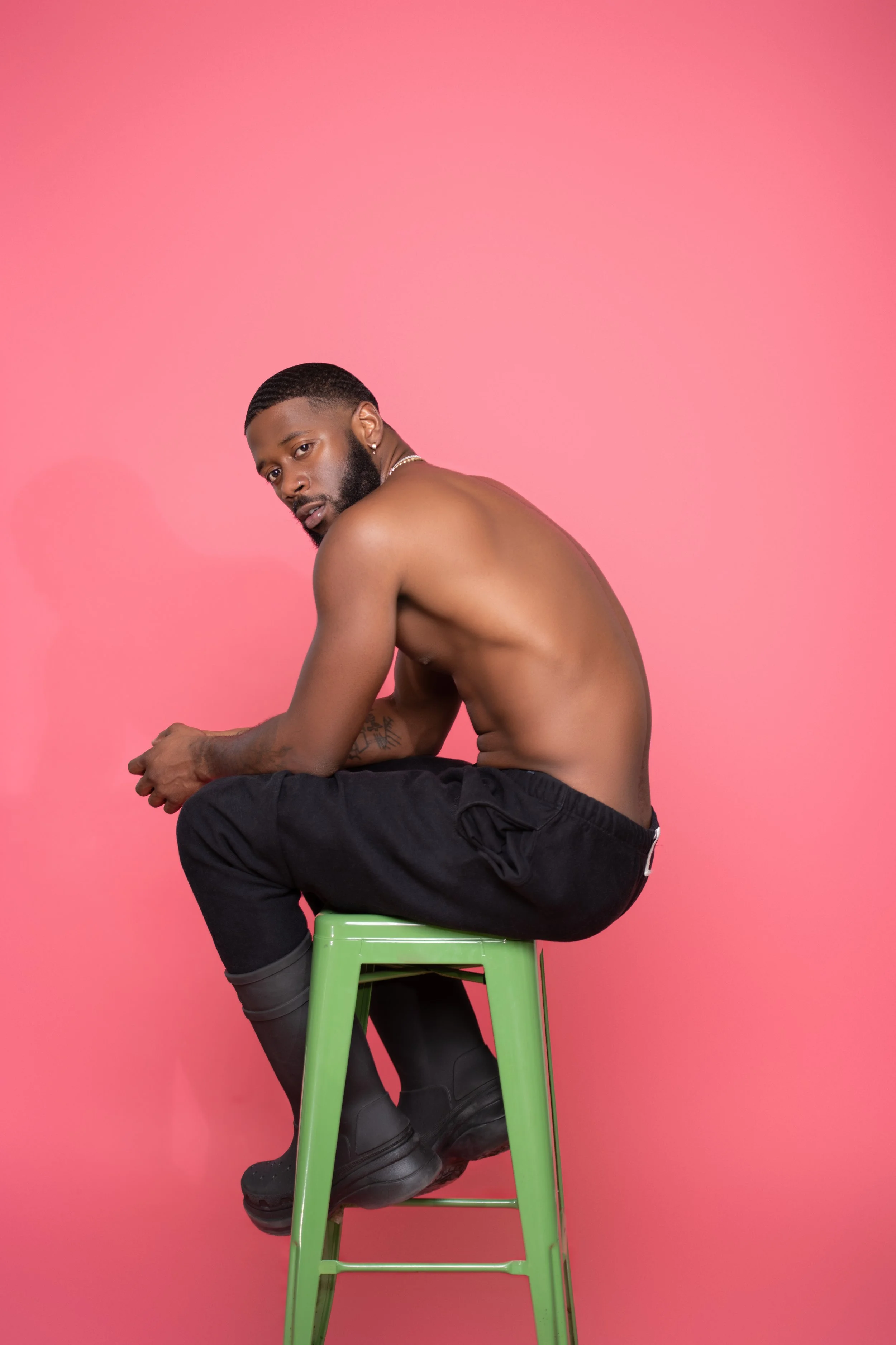 A shirtless man with a beard and earrings is sitting on a green stool against a pink background. He is wearing black pants and black boots, with tattoos visible on his arm. He is sitting with his knees bent and arms resting on his knees, looking at the camera.
