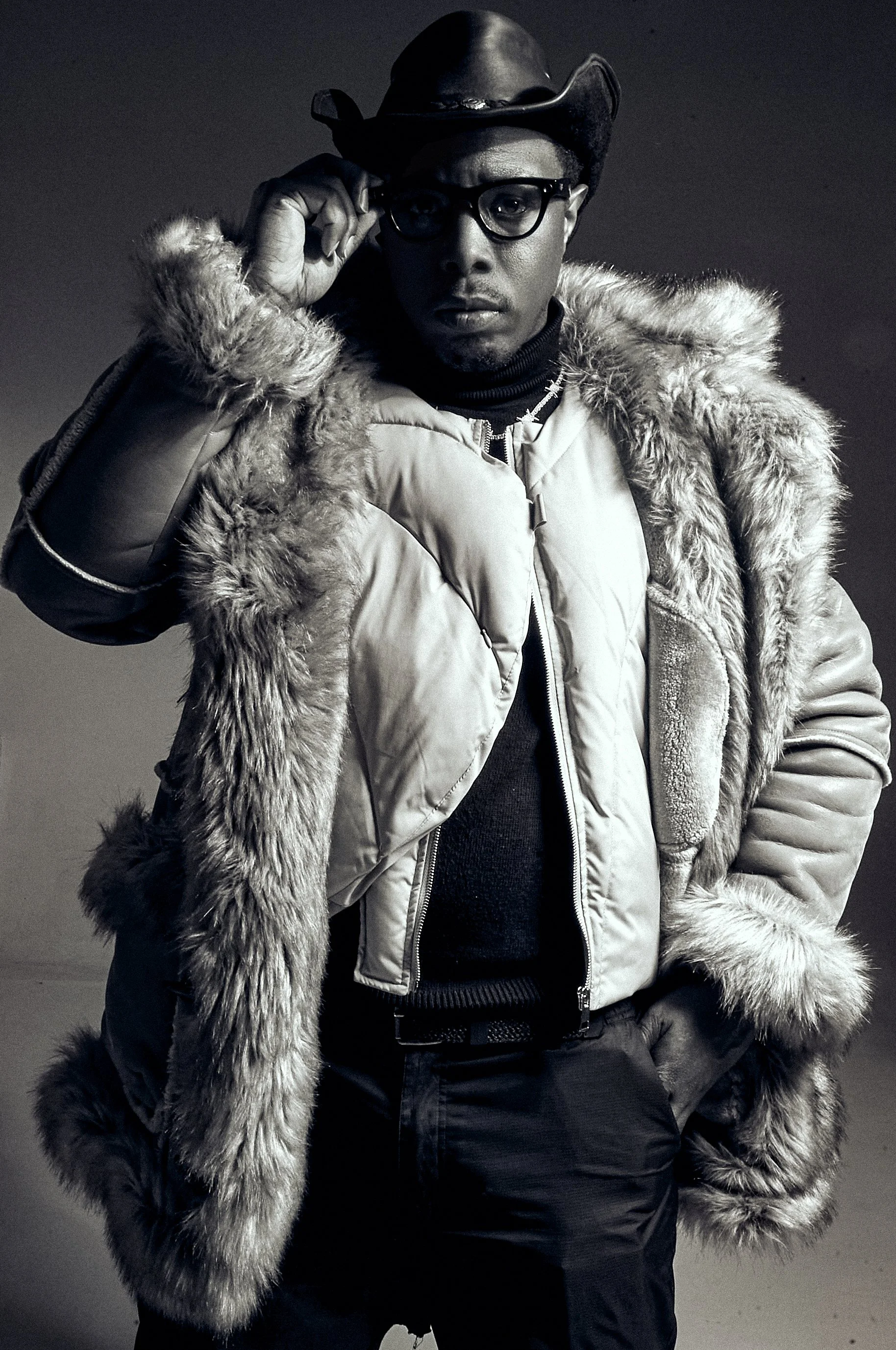A man in a leather cowboy hat, glasses, a fur coat, and a dark turtleneck sweater, posing with his right hand adjusting his glasses in front of a plain dark background.