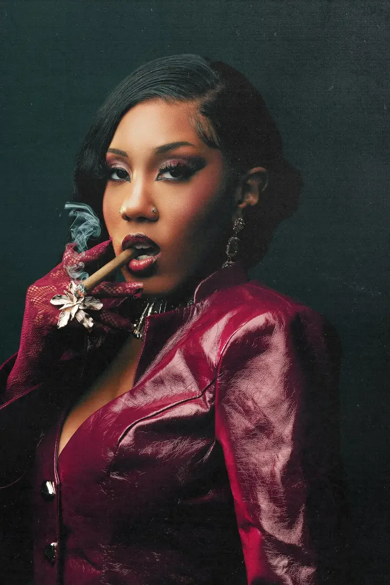 A woman with dark hair styled in waves, wearing a shiny maroon leather jacket, earrings, and lipstick, holding a lit cigar with smoke curling from it, against a dark background.