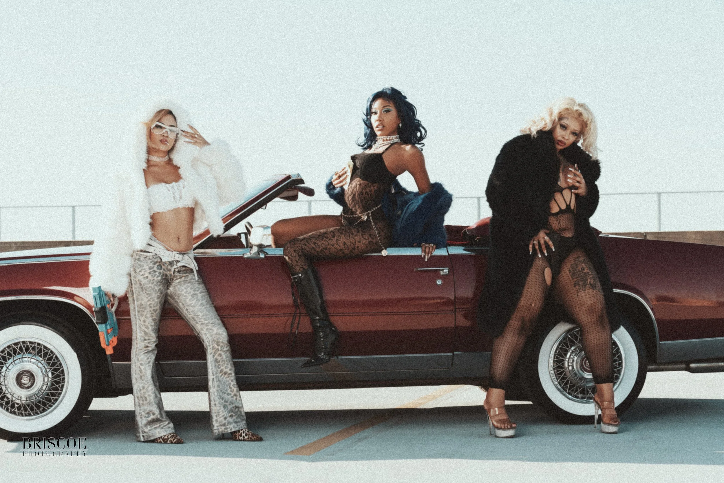 Three women posing by a vintage red car with a parking lot background. One woman is in leopard print pants and a white fluffy top, holding a toy water gun. The second woman is sitting on the car, wearing a sheer animal print dress with a blue jacket and jewelry. The third woman is standing next to the car, dressed in black fishnet stockings and a black fur coat. All are styled glamorously.