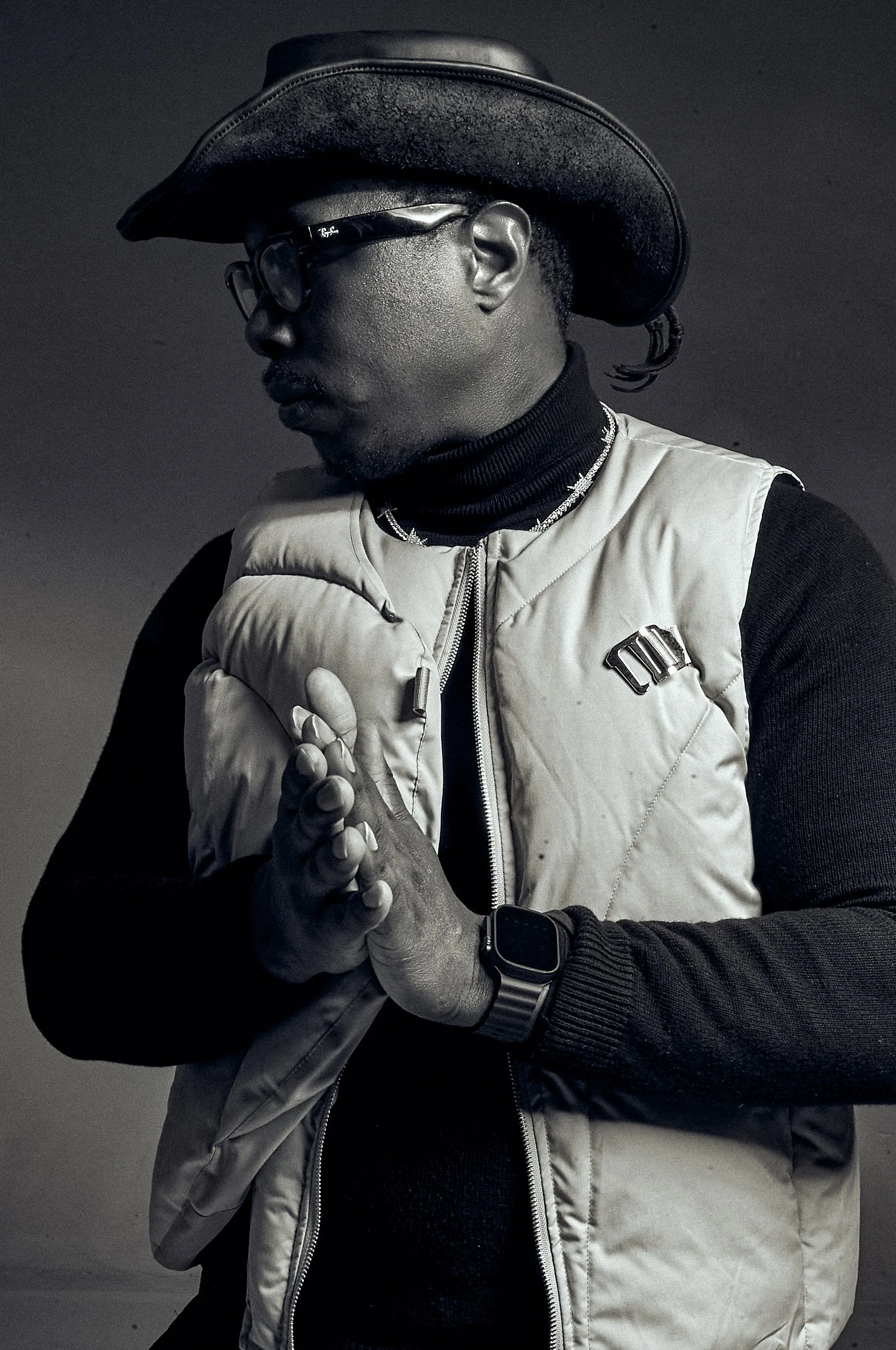A person wearing a cowboy hat, glasses, a black turtleneck, and a beige quilted vest with a 'Dior' logo, standing against a plain dark background with hands pressed together in a prayer-like gesture.