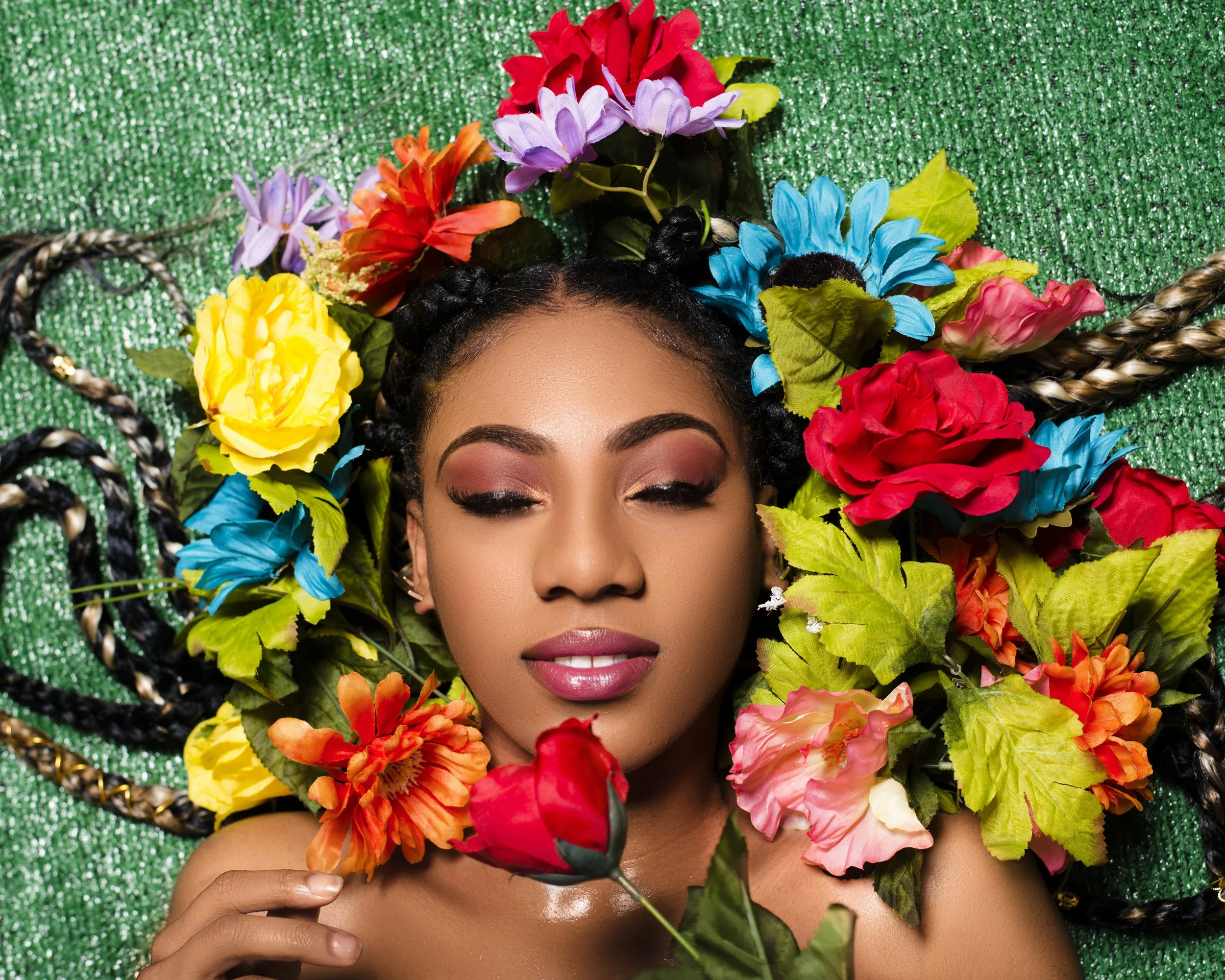 A young woman with braided hair, makeup, and pink lipstick is lying on a green, glittery surface and holding a red rose. She is surrounded by an array of colorful artificial flowers, including yellow, purple, red, pink, orange, and blue, arranged around her head, creating a floral halo effect.