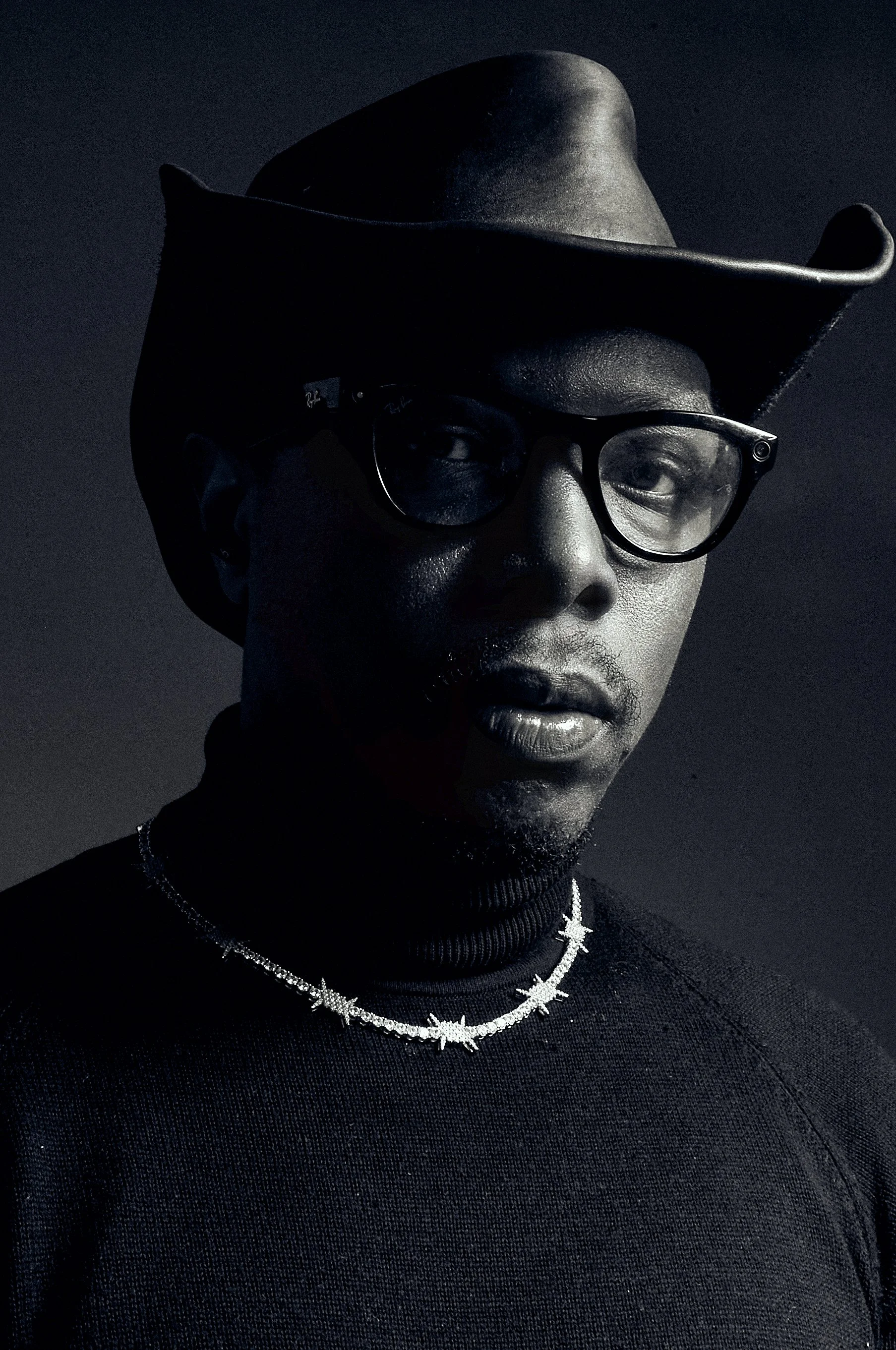 A portrait of a man wearing a black cowboy hat, sunglasses, a black turtleneck, and jewelry including a silver cross necklace and a chain with star-shaped pendants. The photo is in black and white.
