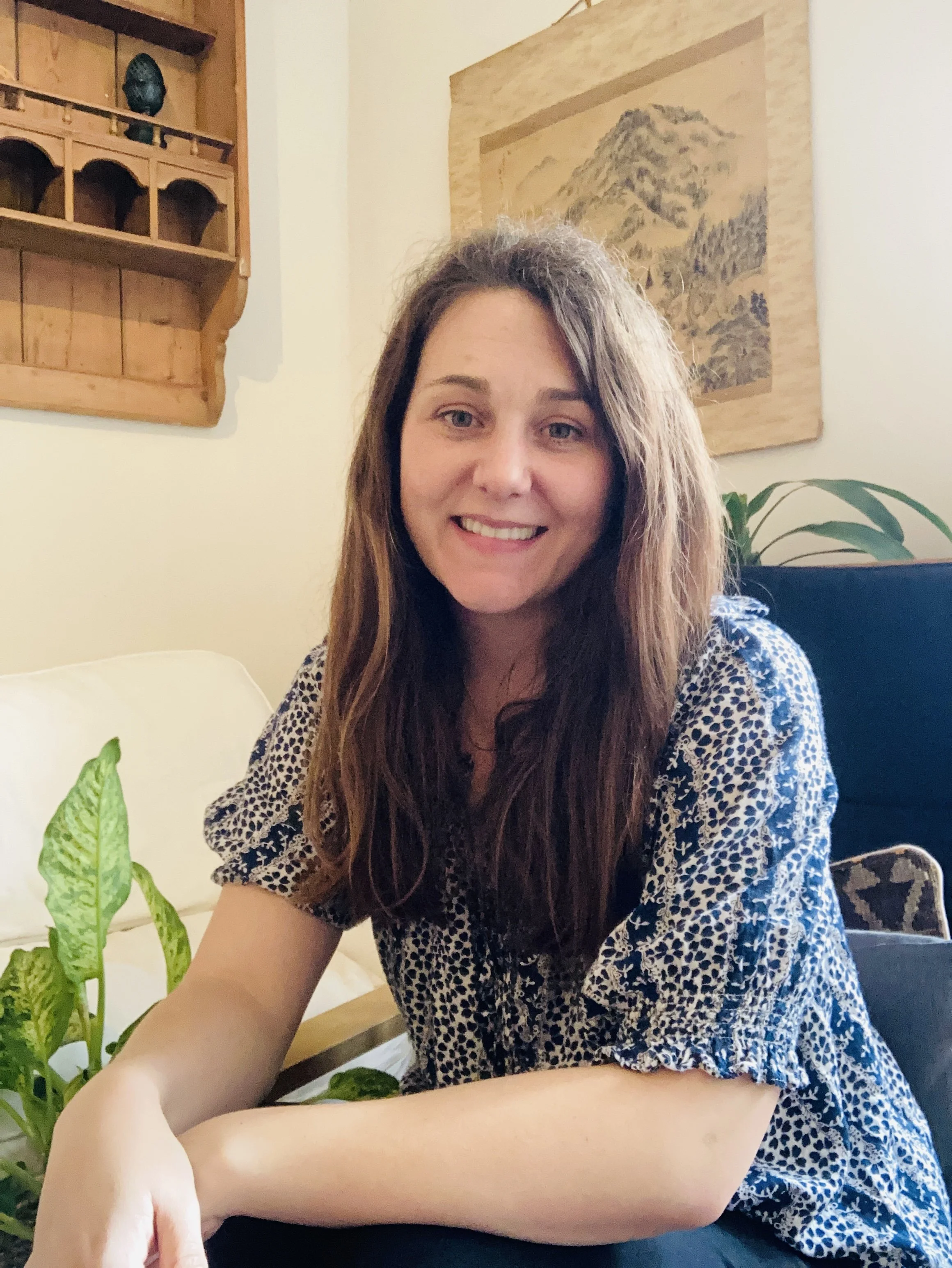 Hannah, a Counsellor with long brown hair smiling, sitting in a welcoming cozy therapy room. In the background, there's green plants and pictures and a wooden shelf with decorative items.