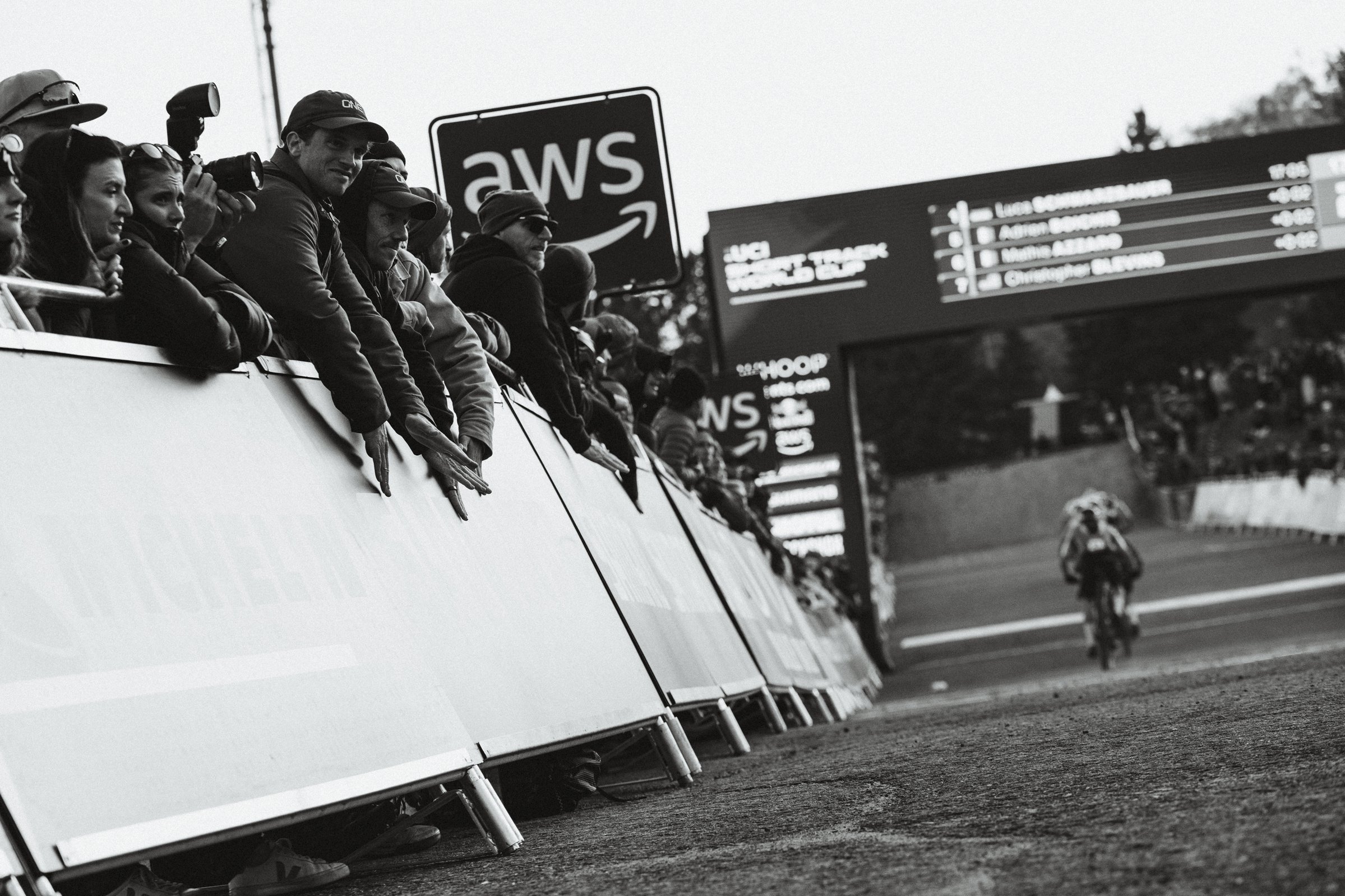 Spectateurs regardant une course de cyclisme au bord de la route avec une affiche de l'événement et un écran de classement en arrière-plan.