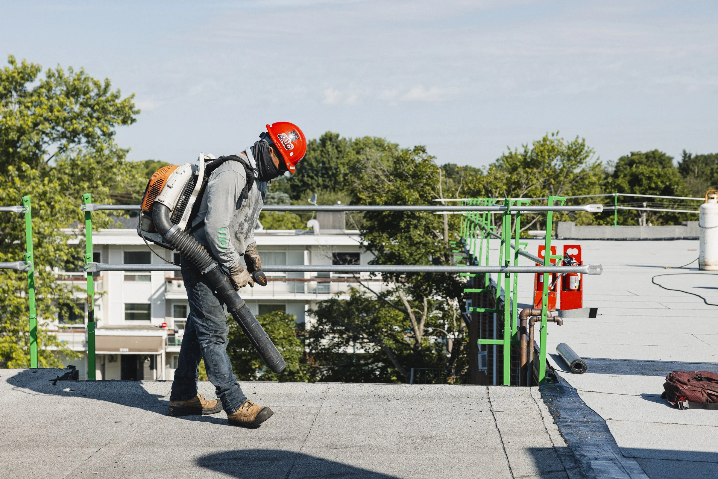 Un ouvrier portant un casque rouge et un masque utilise un aspirateur pour enlever la poussière ou les débris sur le toit d’un bâtiment en construction.
