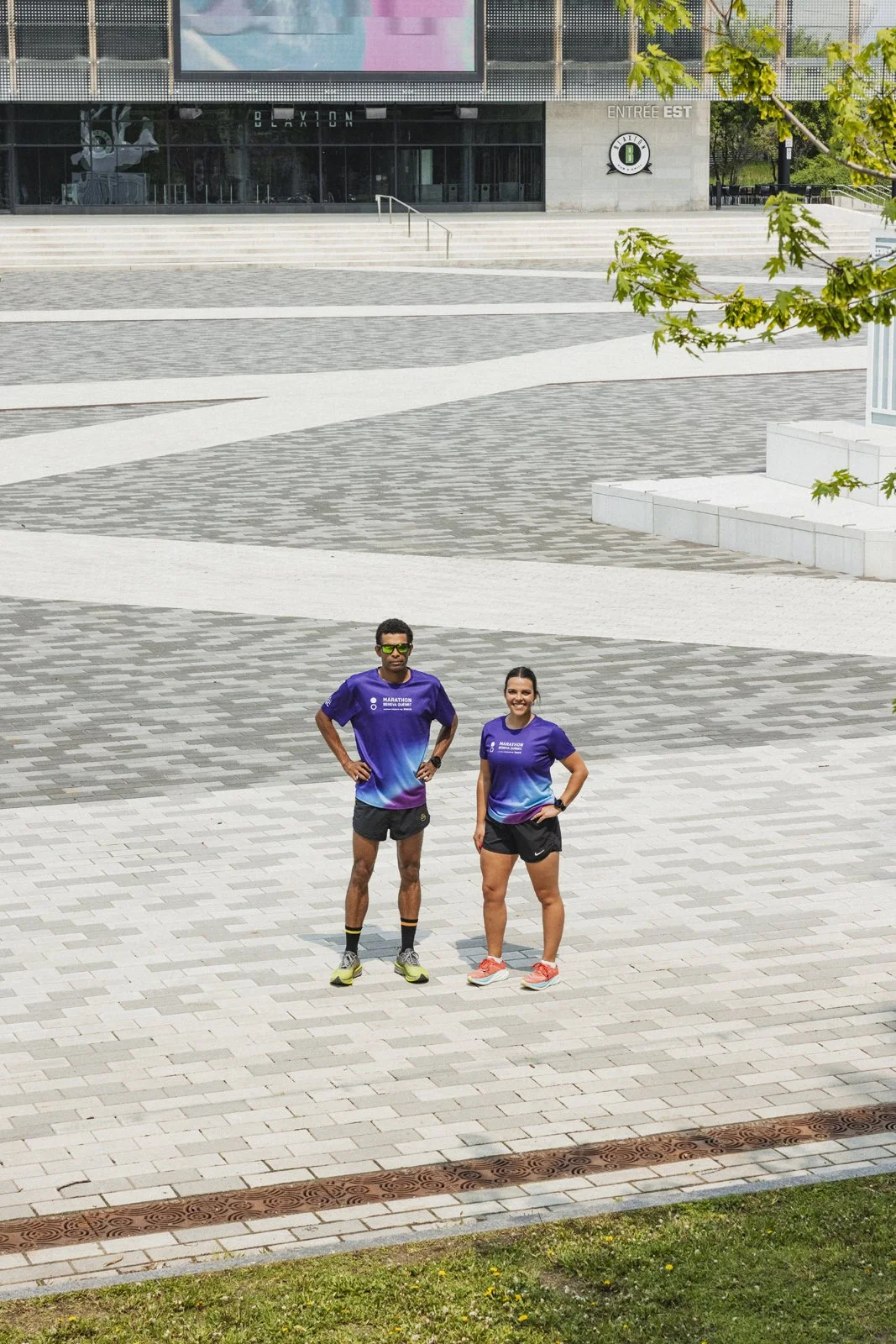 Deux coureurs en tenues violettes posent sur une place pavée devant un bâtiment moderne avec un grand écran et un panneau indiquant 'ENTRÉE EST'.