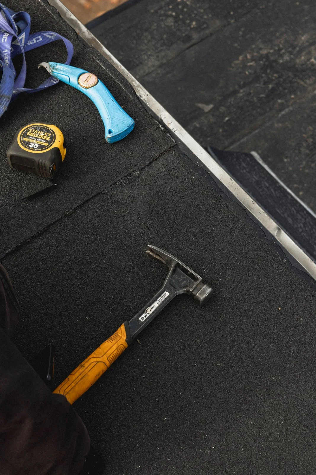 Un marteau avec une tête en métal et un manche en bois, une clé à tête plate, un ruban à mesurer de marque Stanley, et un tournevis électrique bleu sur une surface noire de toiture.