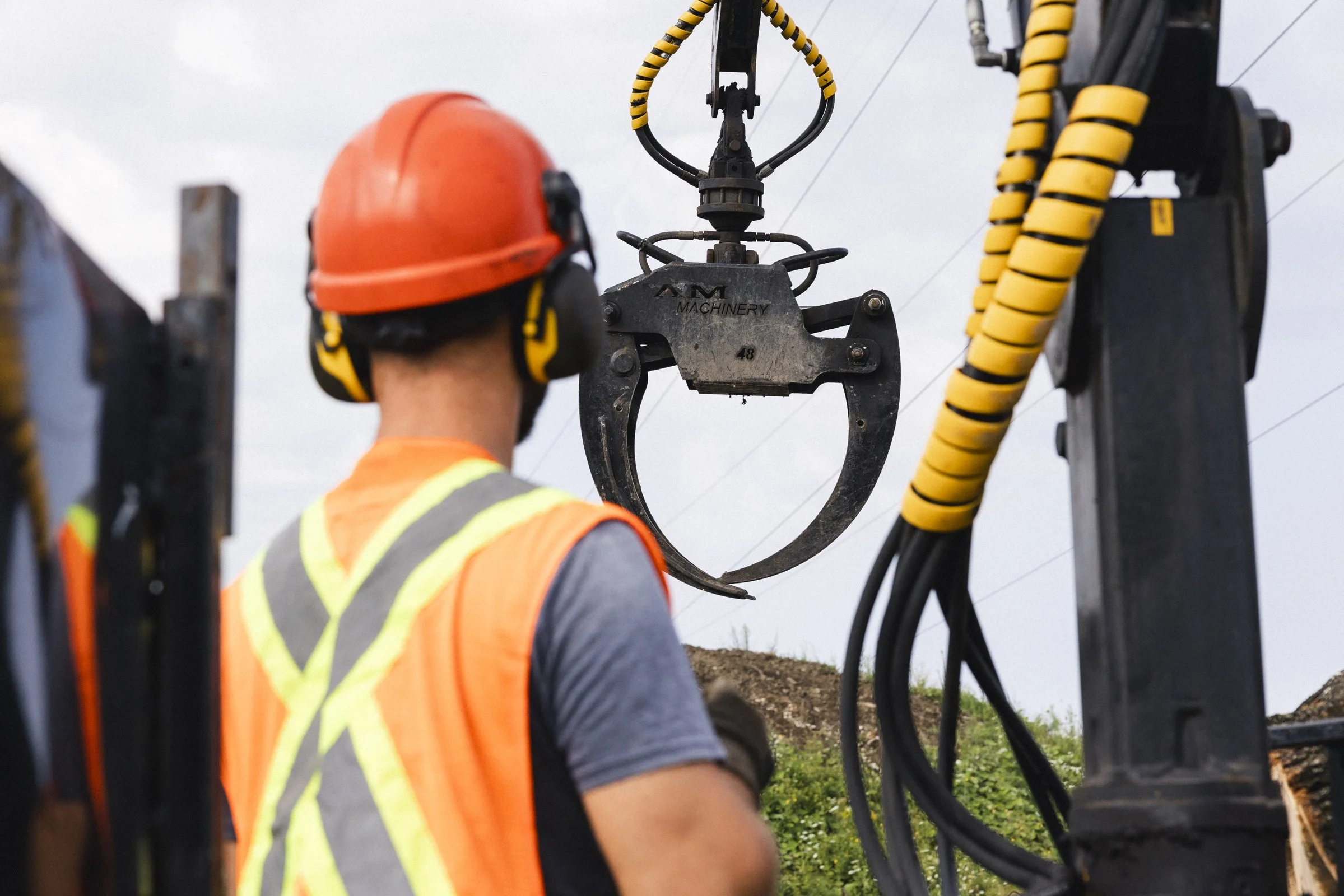 Ouvrier portant un casque de sécurité orange et un gilet réfléchissant à côté d'une machinerie avec une pince géante, dans un environnement extérieur.