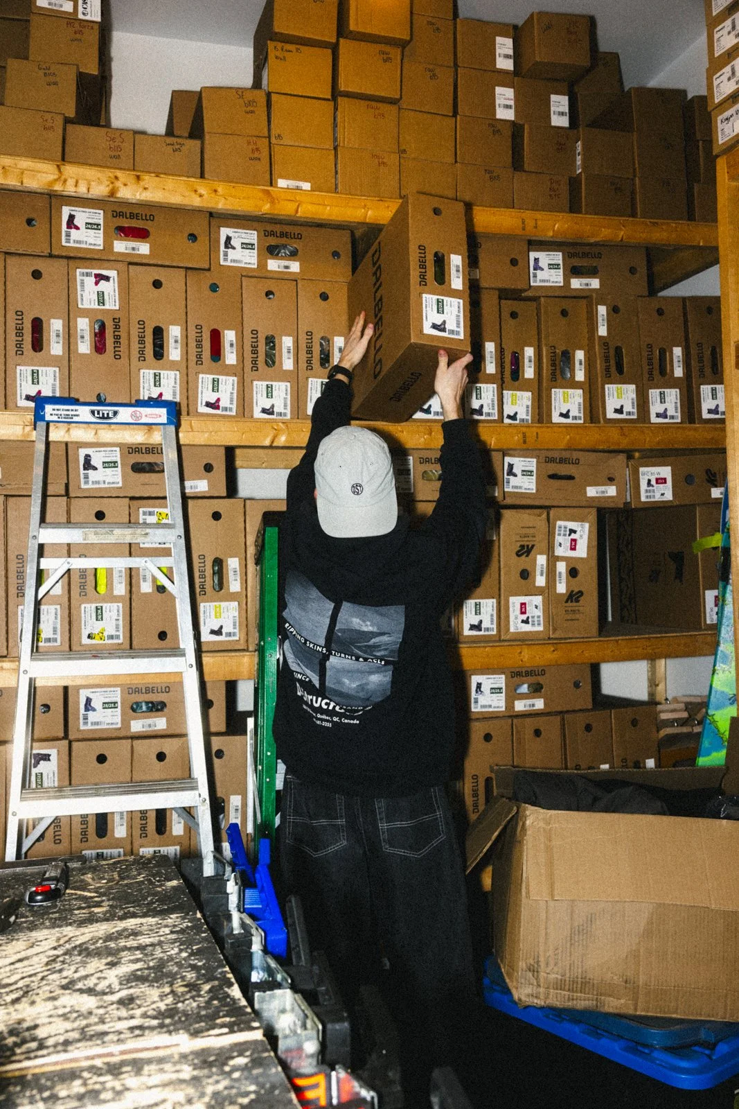 Une personne en noir avec un chapeau gris organise des boîtes en carton sur des étagères dans un entrepôt.