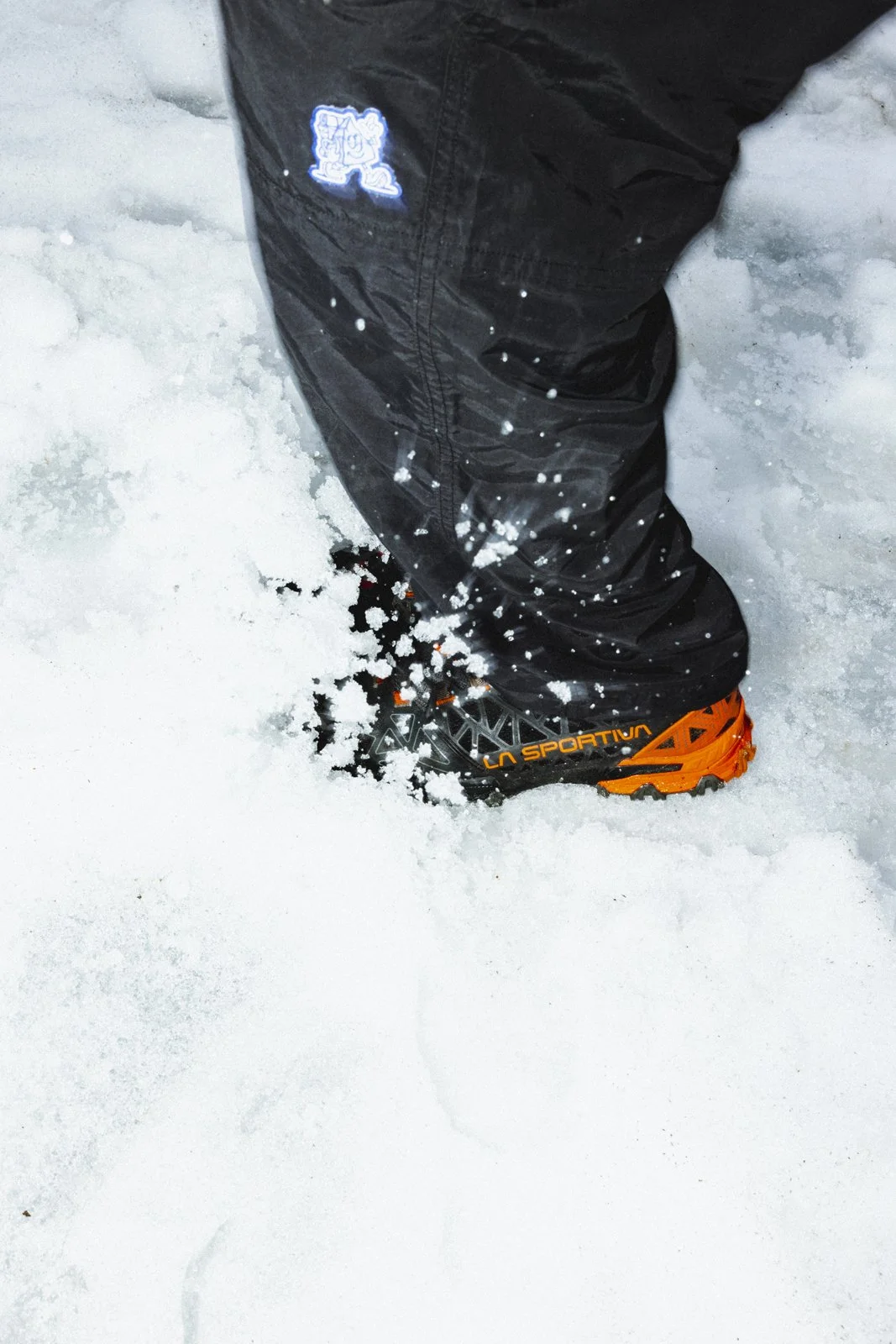 Une personne en bottes de randonnée orange et pantalons noirs marche dans la neige, laissant des empreintes dans la neige fraîche.
