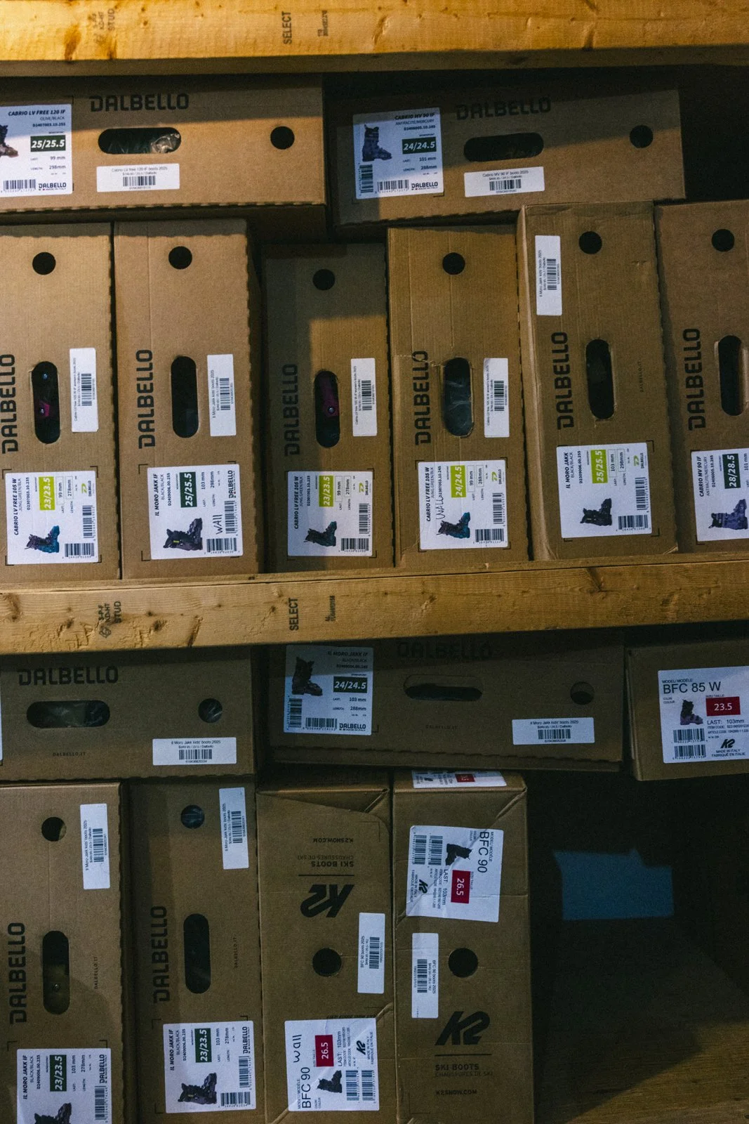 Étagère avec des boîtes en carton contenant des bottes de ski Dakbello, avec différentes tailles et étiquettes visibles.