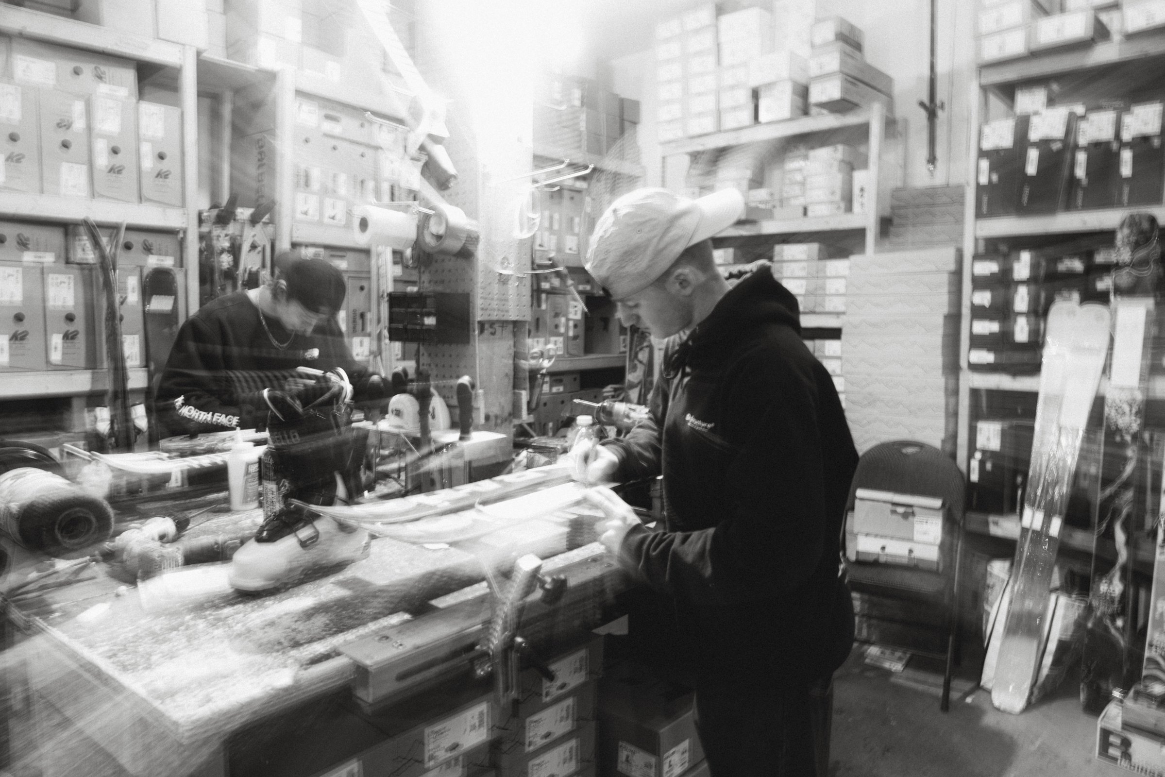 Deux jeunes personnes travaillent dans un atelier ou un magasin, entourées de matériel et de boîtes sur des étagères.