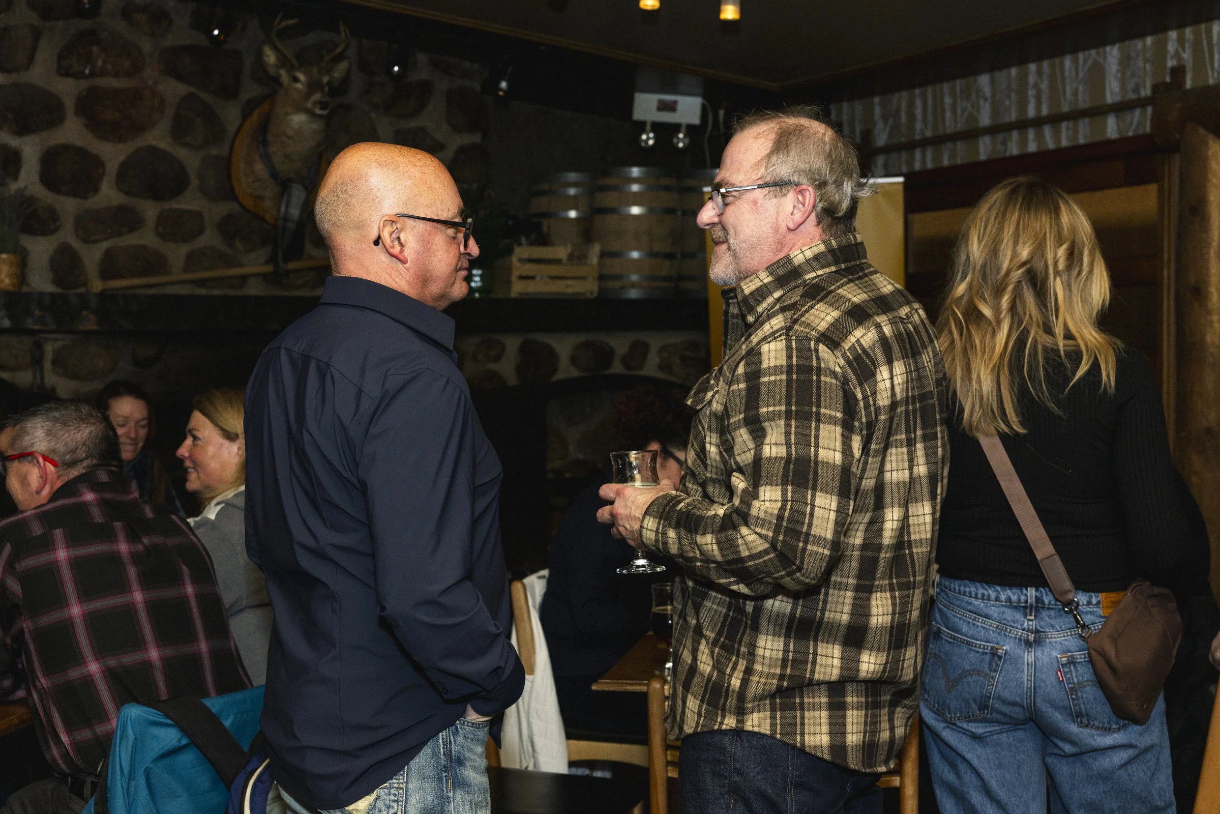 Deux hommes discutent dans une ambiance chaleureuse, tenant des verres, dans un restaurant ou un pub avec un mur en pierre et un trophée de cerf en arrière-plan.