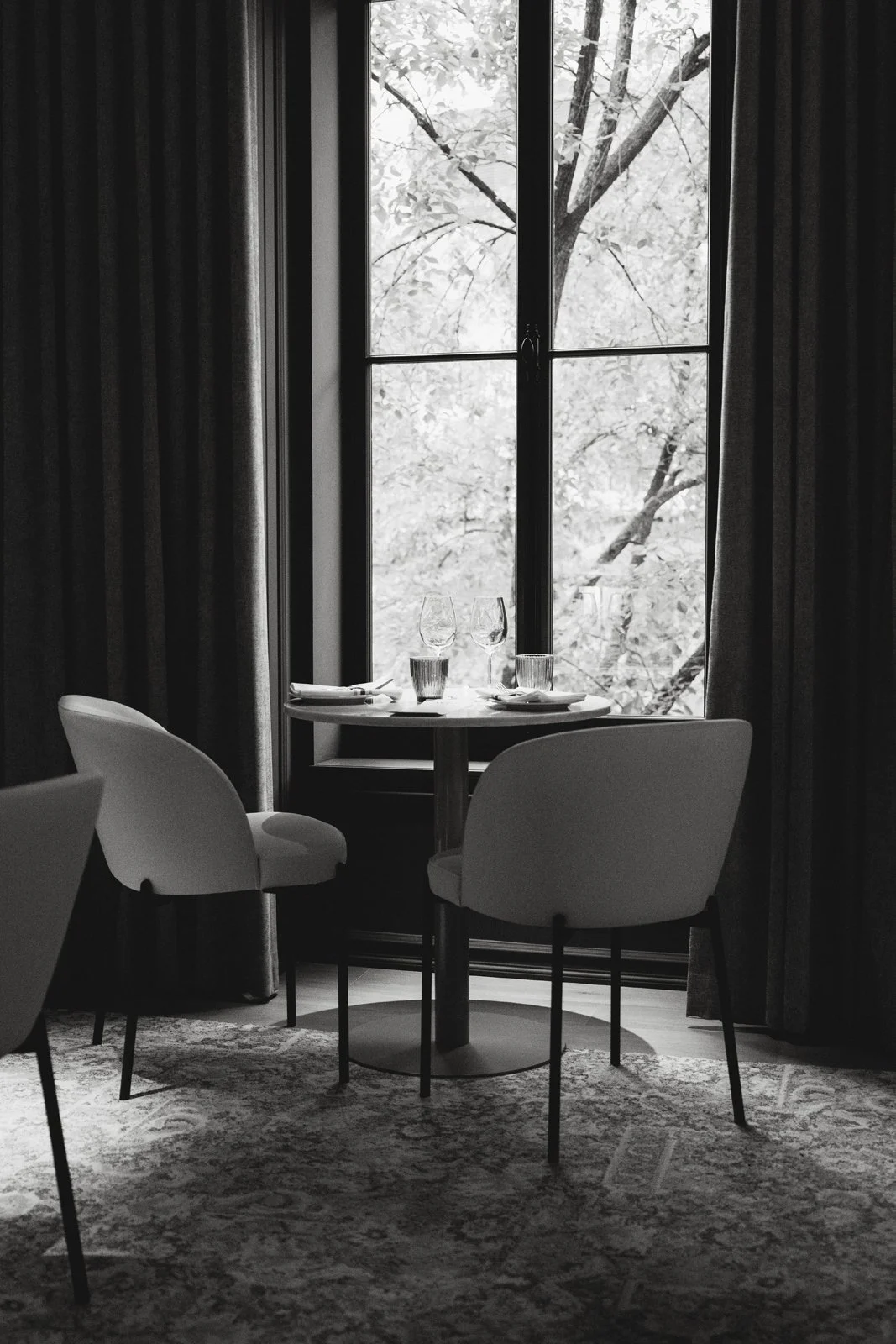 Une table de restaurant élégante près d'une grande fenêtre avec vue sur des arbres, décorée avec deux verres à vin, deux verres d'eau, et des assiettes, dans une ambiance monochrome.