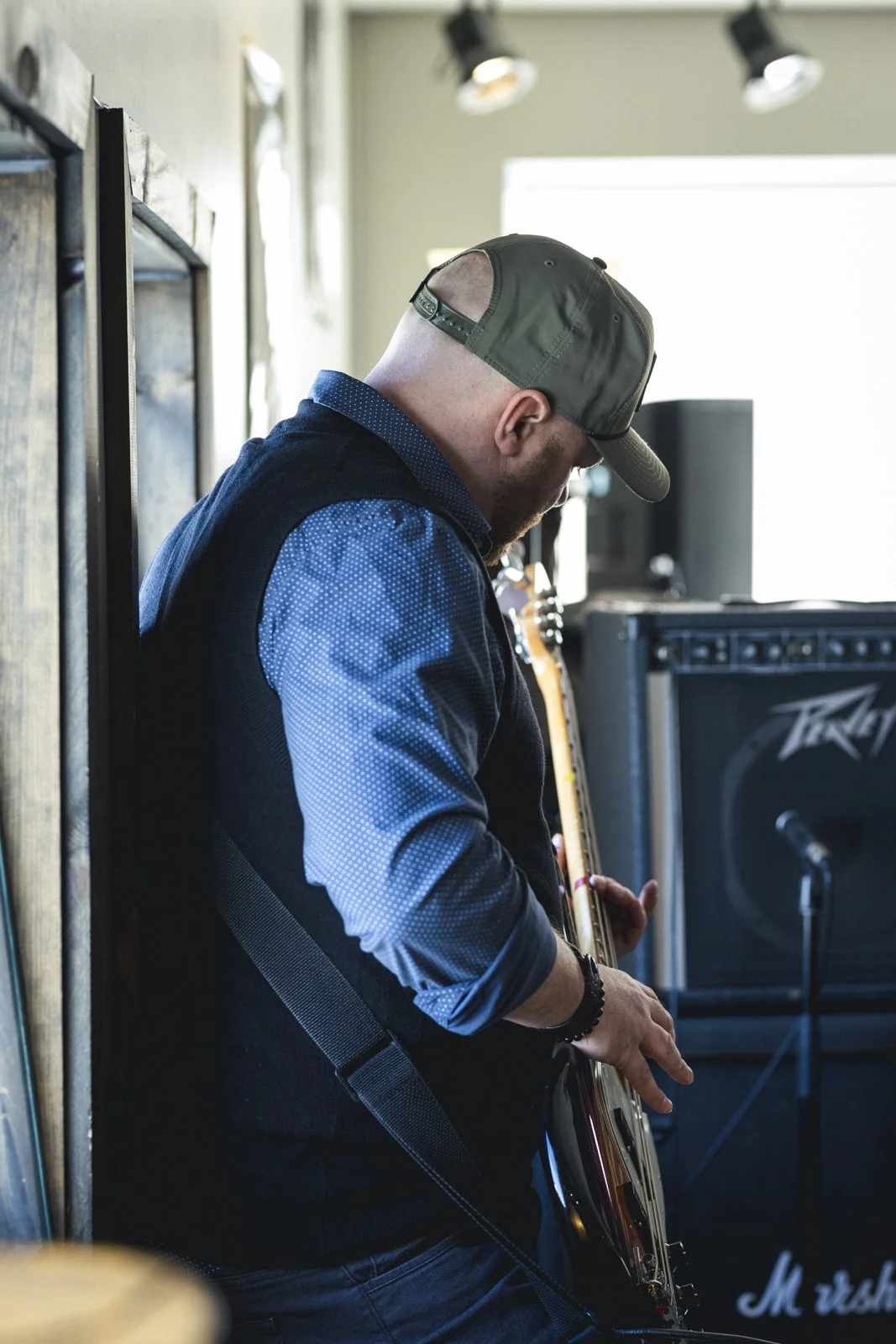 Jeune homme jouant de la guitare électrique dans une salle de musique. Il porte une casquette noire, un sac à dos noir et une chemise à pois bleus. En arrière-plan, une amplificateur de musique et un microphone.