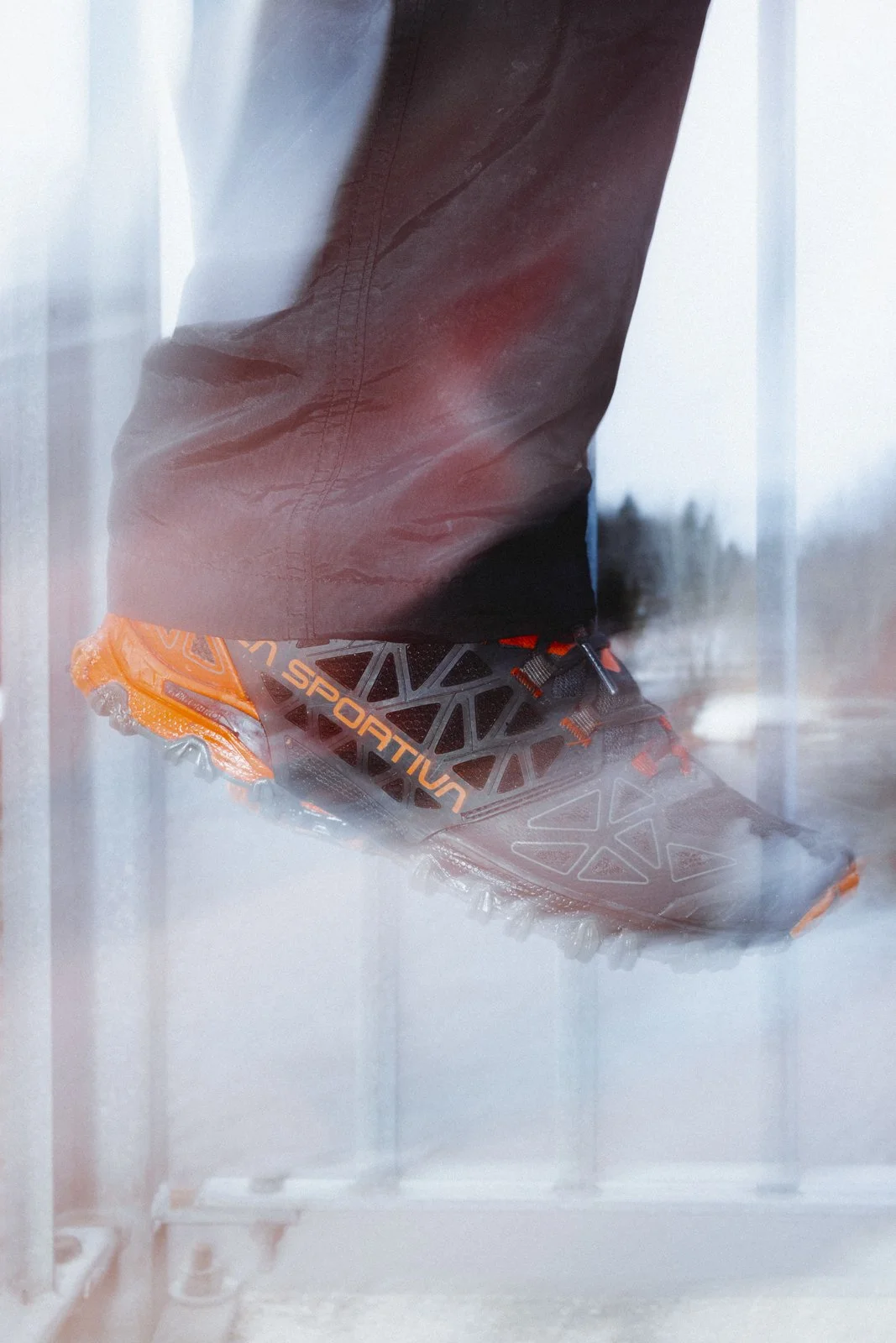 Une chaussure de trail running orange et noire avec la marque 'Sportiva', portée par une personne en pantalon noir, vue à travers une vitre ou un plastique.