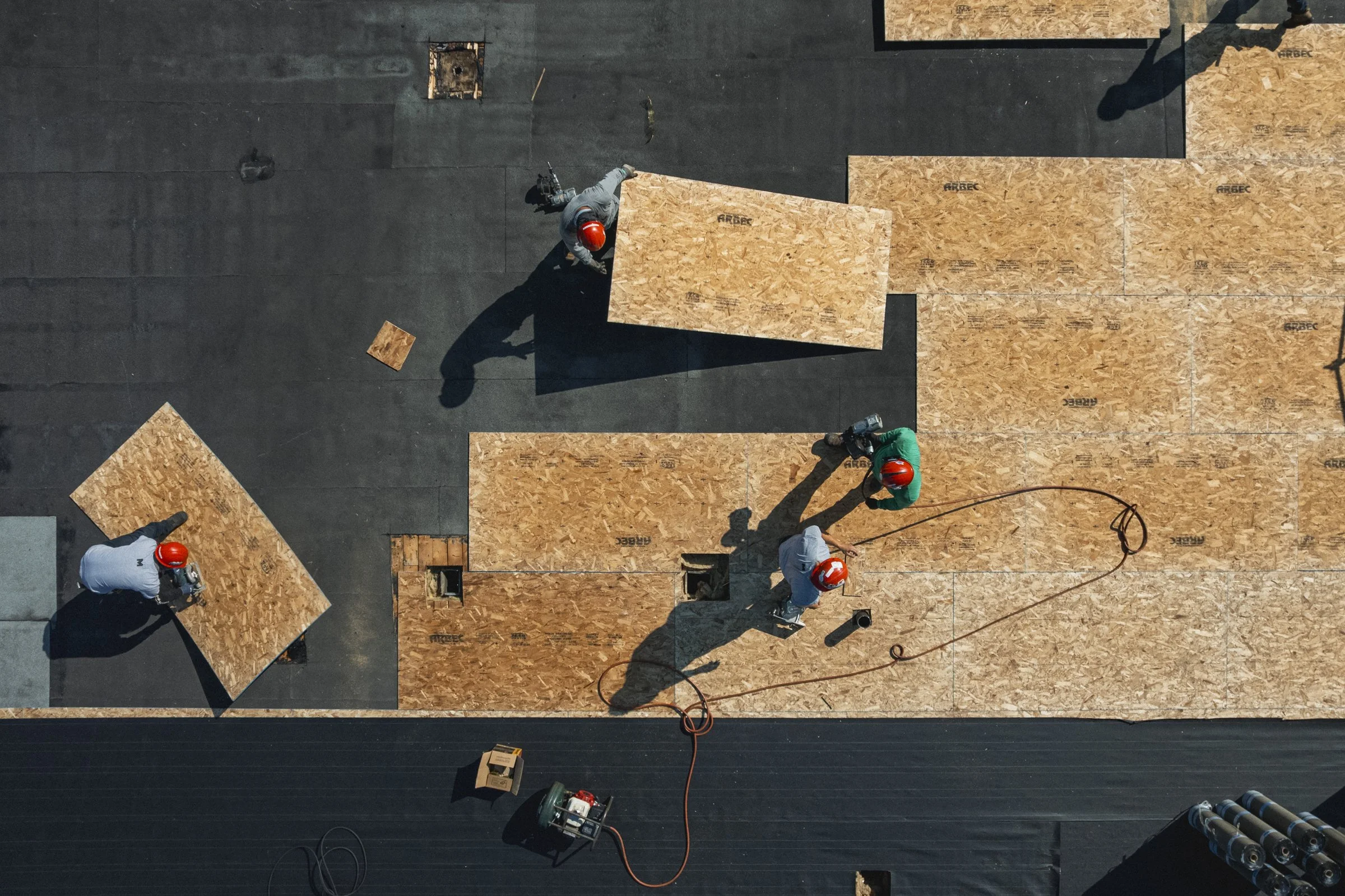 Travail en équipe, cinq travailleurs portant des casques rouges, installant des panneaux de bois sur un toit en construction, avec outils et câbles électriques autour.