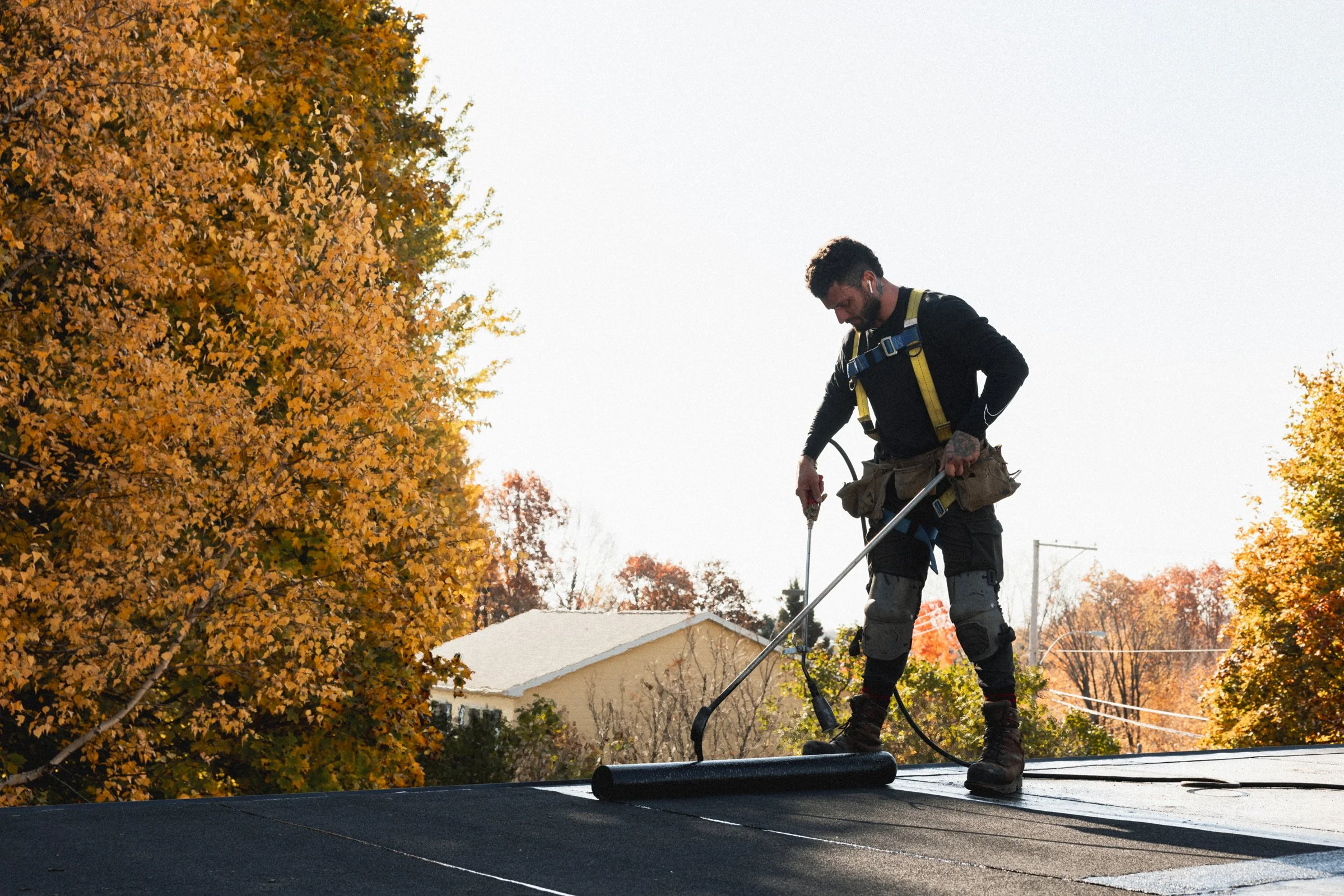 Un homme en tenue de travail pose un revêtement sur un toit en utilisant une rouleau, entouré d'arbres aux feuilles d'automne en arrière-plan.