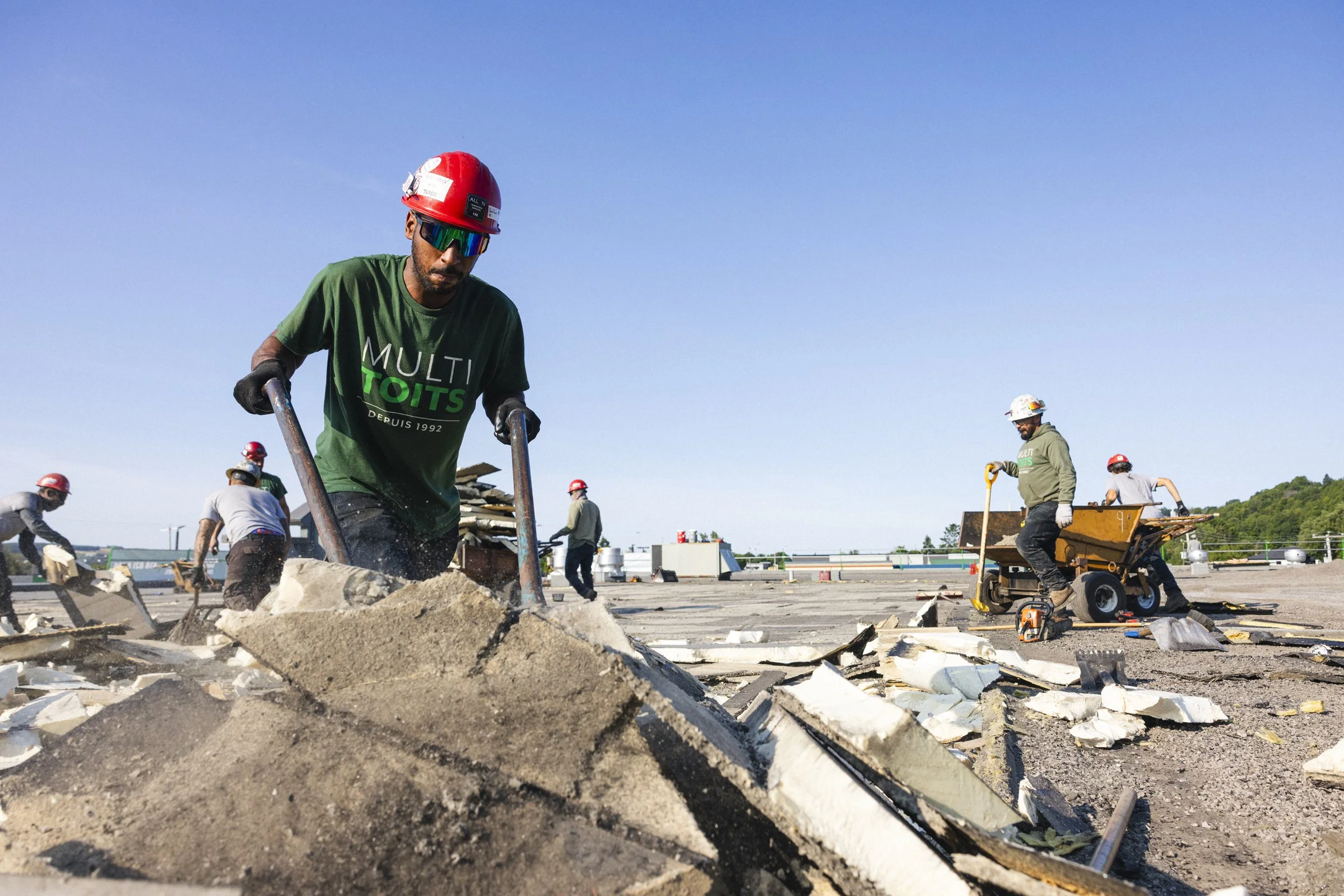 Travailleurs portant des casques de sécurité et des gants, nettoyant et préparant un toit en construction avec des outils, dans un environnement industriel en plein air par une journée ensoleillée.